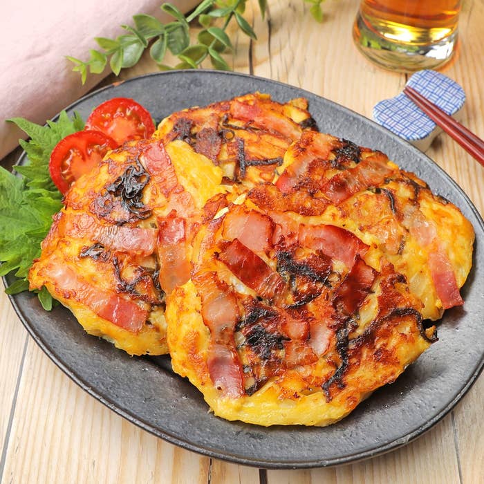 黒い皿に、ベーコンと野菜が入ったお好み焼きが盛られている。横にトマトのスライスが添えられている。