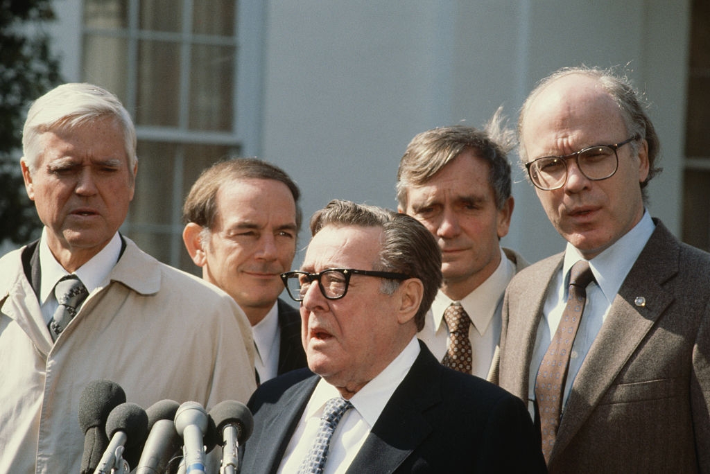 Five men in suits and coats stand before microphones outside a building, appearing to address the media during a press event