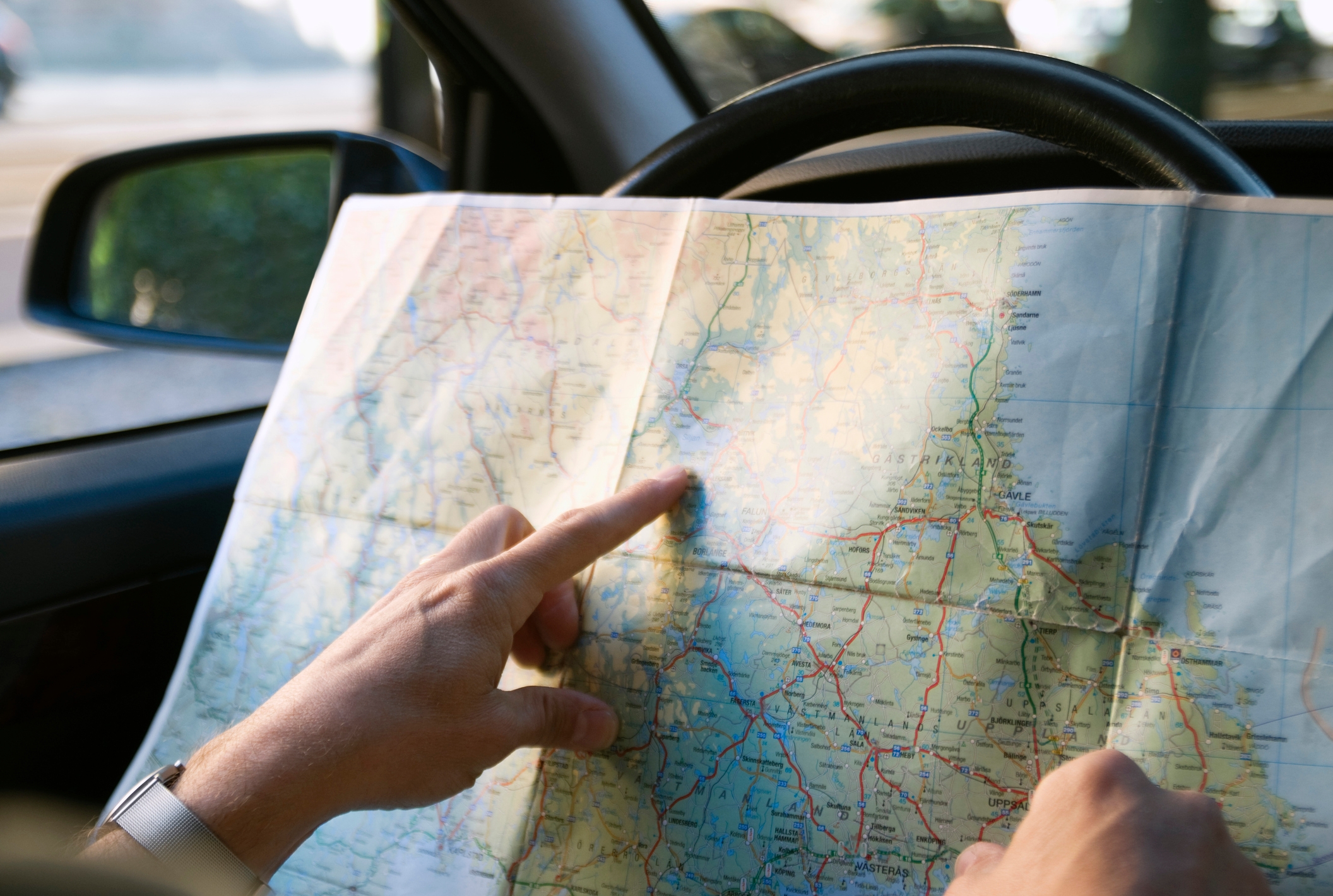 Person in car holding and pointing at a road map, showing a journey or navigation scene. Steering wheel is visible