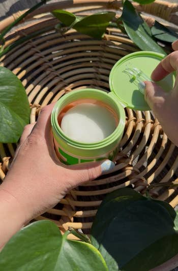 reviewer opening a skincare jar with a toner pads inside, surrounded by greenery connected a woven tray