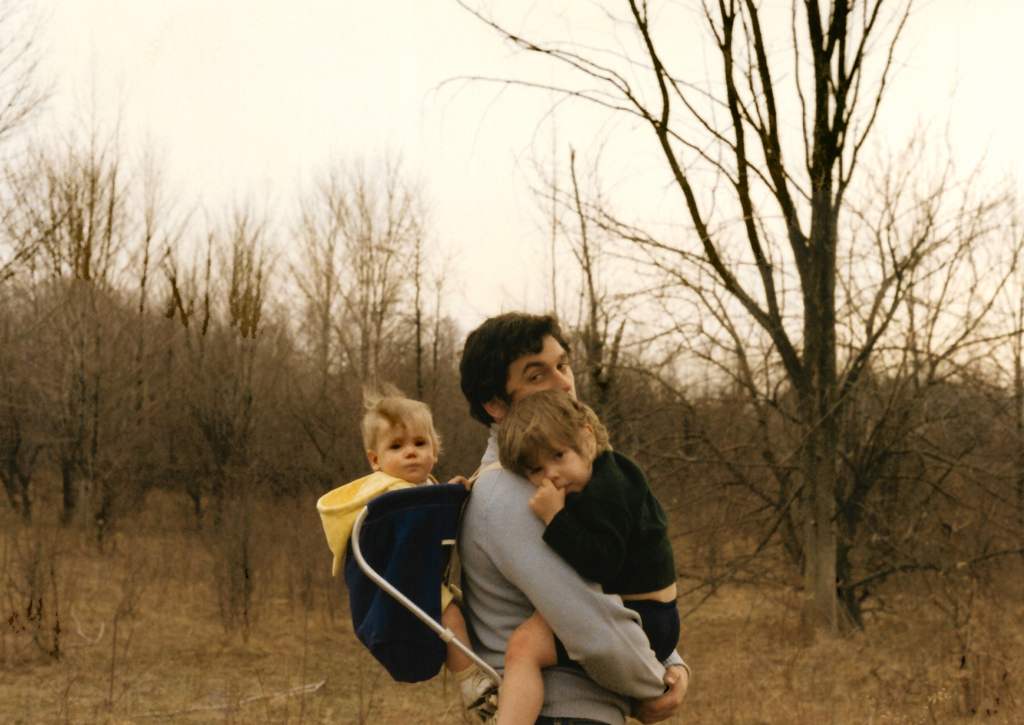 A man carries two young children, one in a backpack carrier, while walking through a leafless forest area