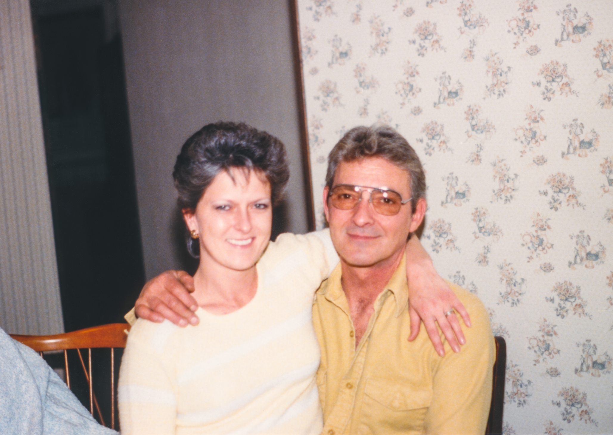 A smiling man in glasses and a yellow shirt sits with a woman in a striped sweater, their arms around each other in a casual setting