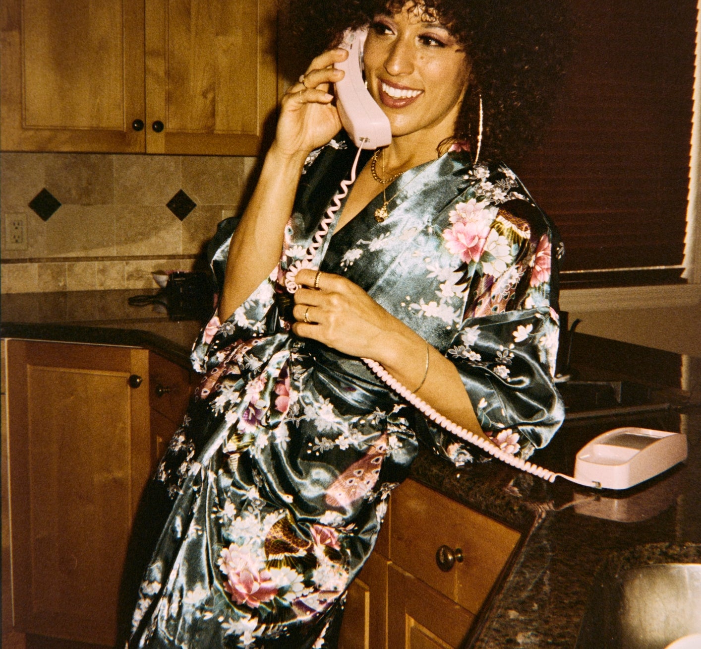 Person in a floral robe smiling while talking on a vintage pink telephone in a kitchen