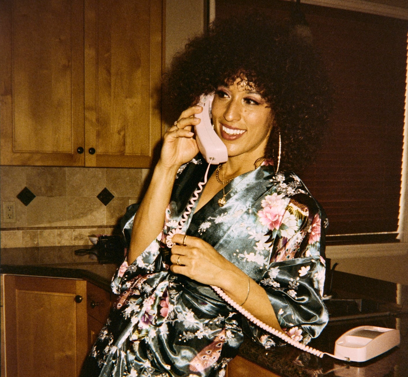 Person in a floral robe smiling while talking on a vintage pink telephone in a kitchen