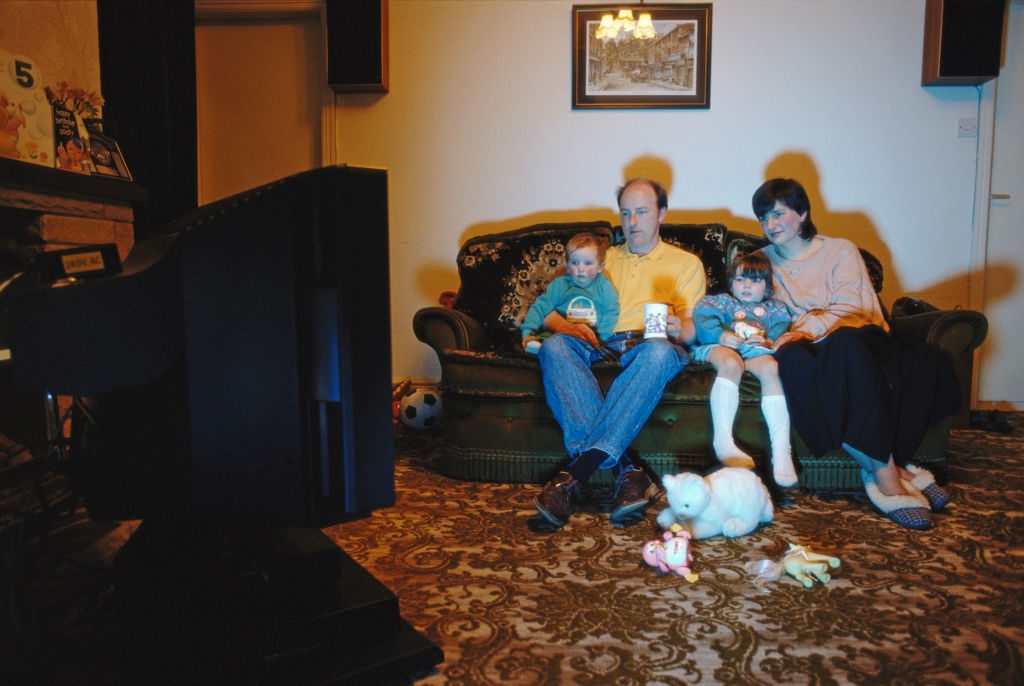 Family watching TV in a cozy living room, surrounded by toys; two children sit between parents on a patterned sofa