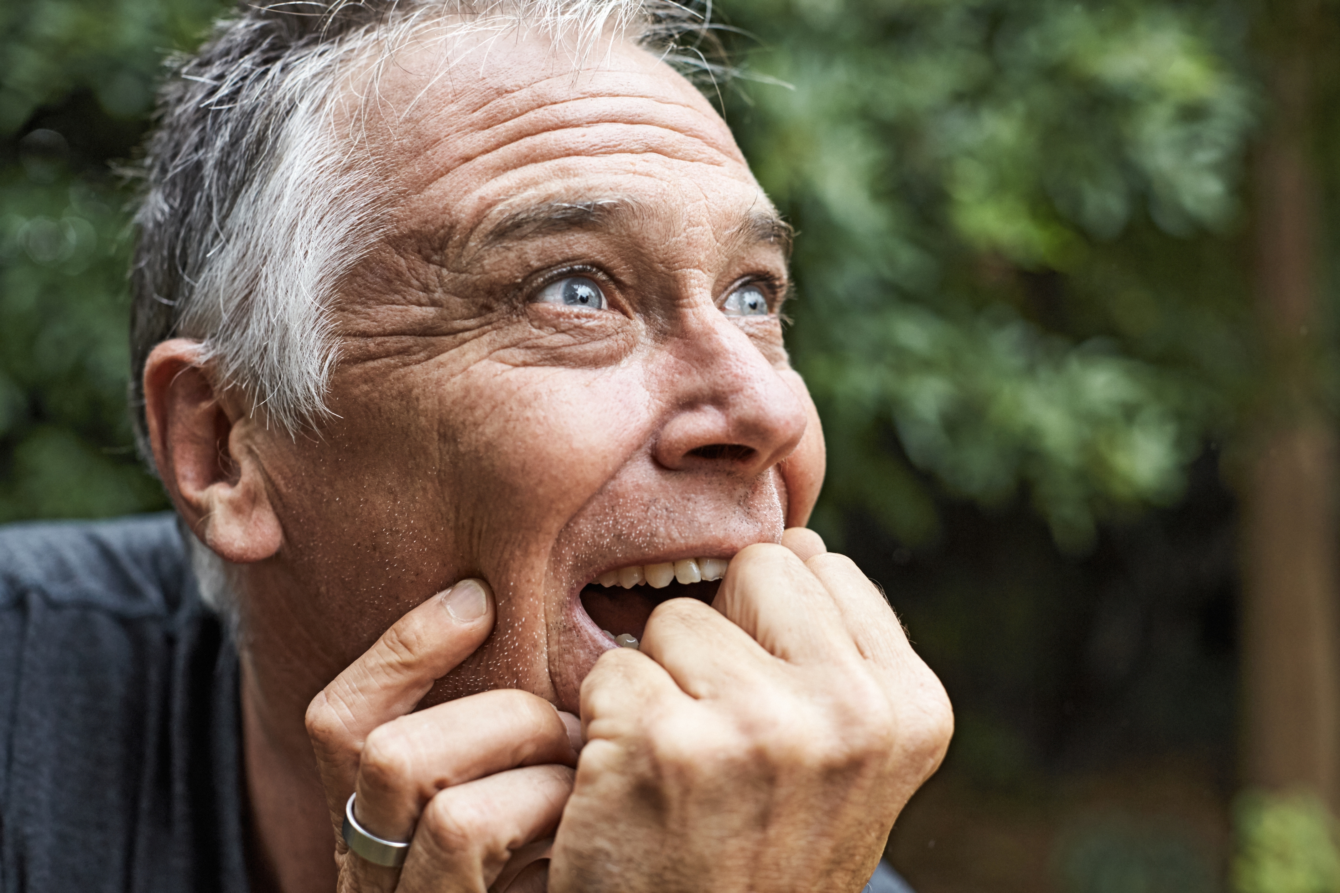 A person with short gray hair looks up with a joyful expression, hands held near the mouth. Blurred greenery is in the background