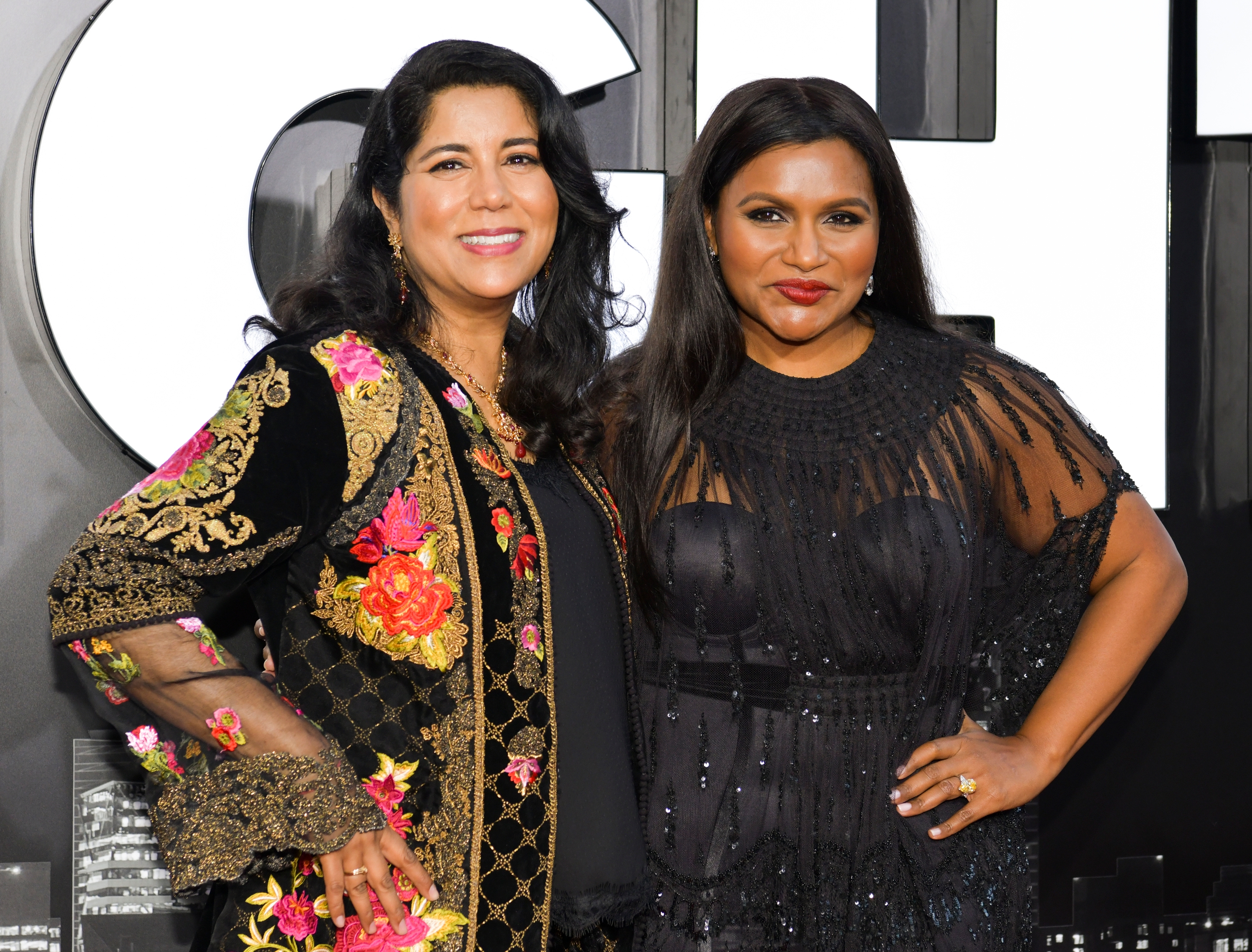 Two women smiling at an event. Left woman wears a floral-patterned jacket, right woman wears an elegant black dress with sheer elements