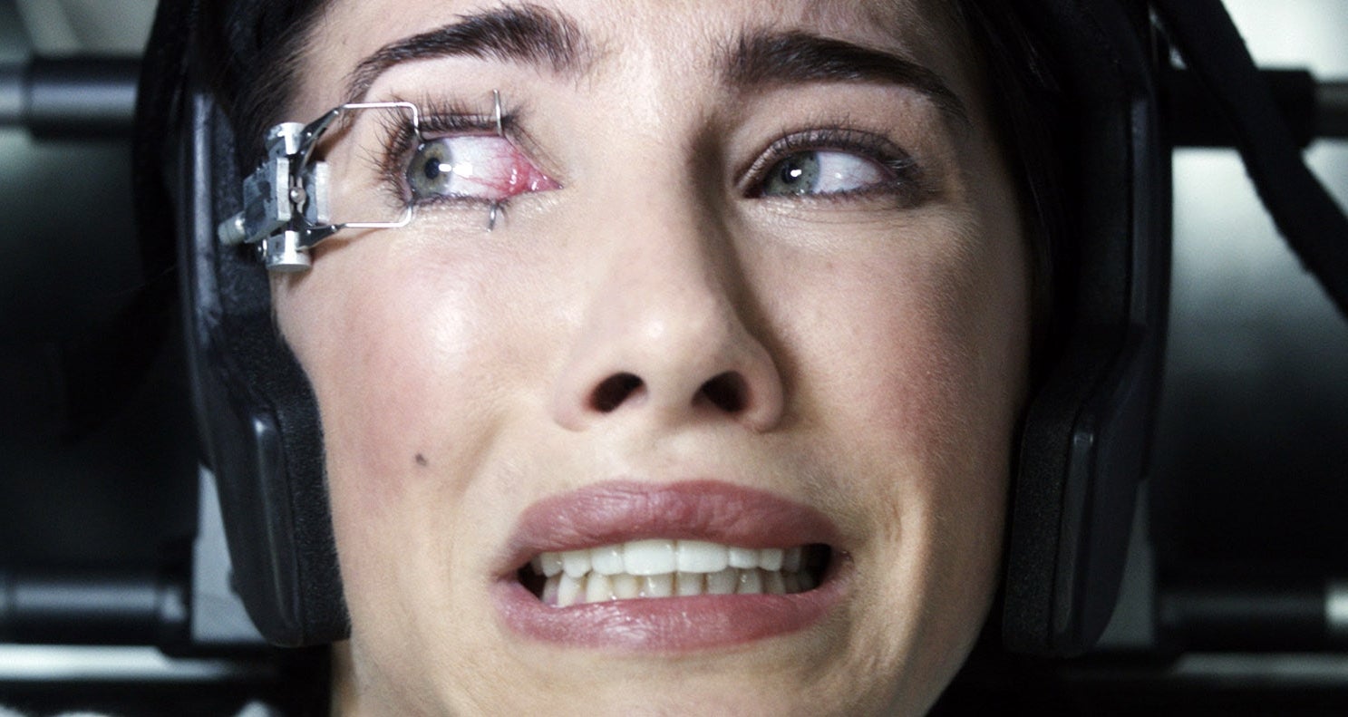 A person with an eye held open by a device looks frightened, lying down with their head supported
