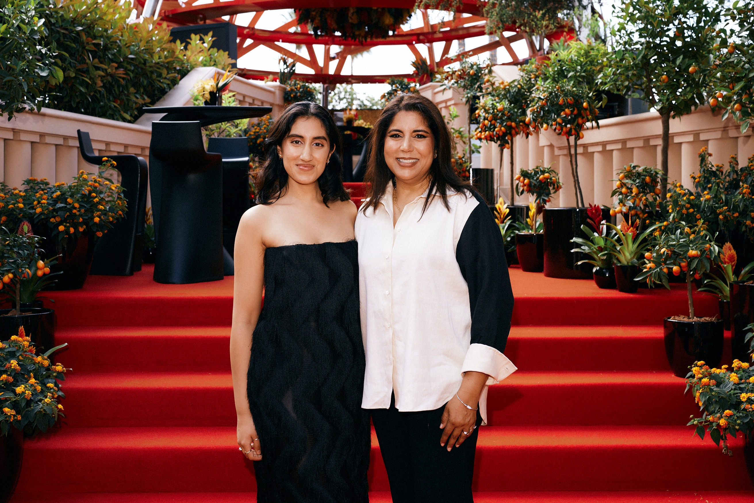 Two people stand on red steps, one in a strapless textured dress, the other in a blouse and pants. Orange plants are arranged around them