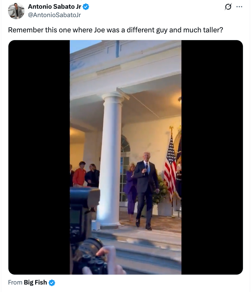A man in a suit speaking behind a podium on a porch with American flags. Photographers are capturing the event