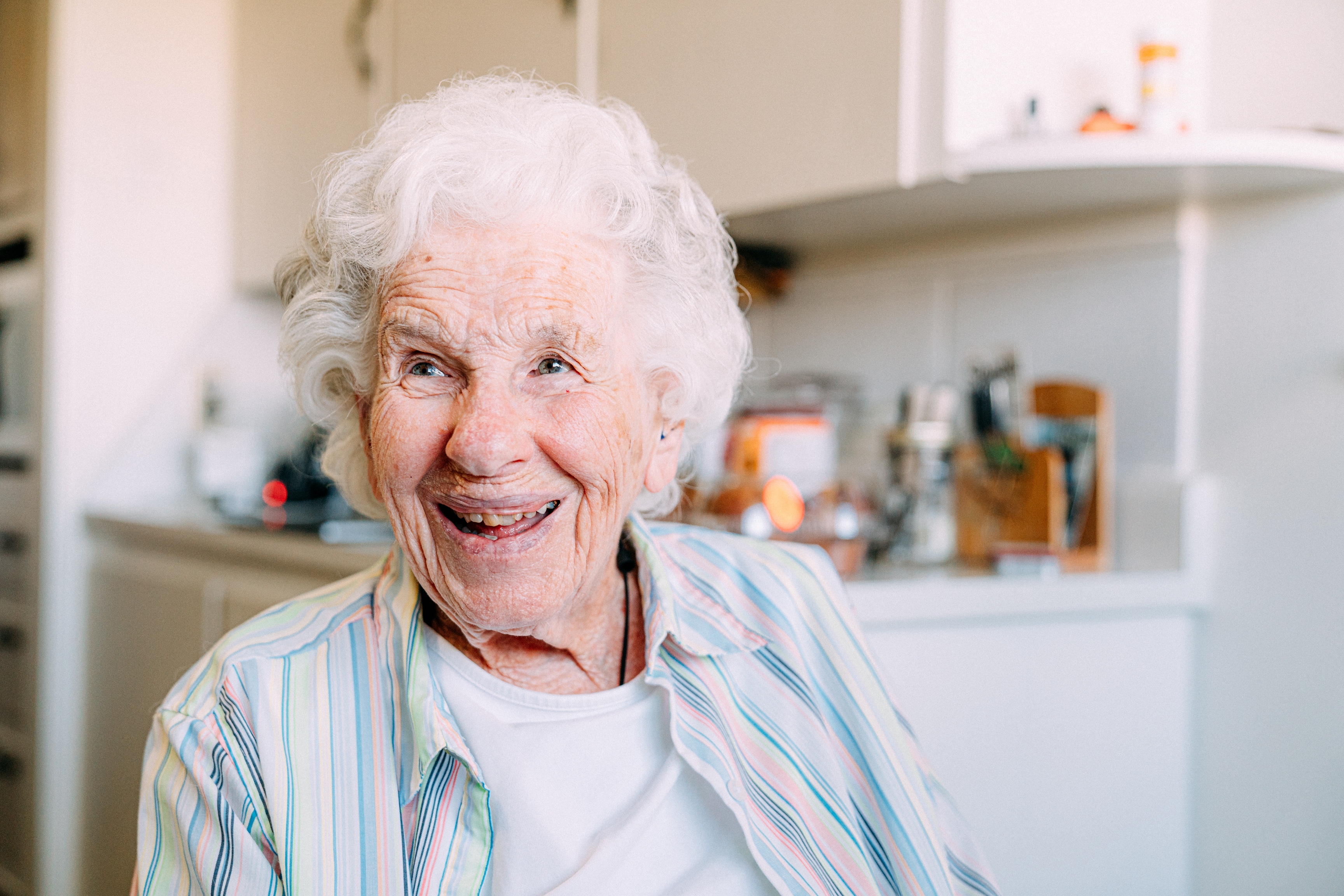 Elderly woman with curly hair, wearing a striped shirt, smiles warmly while sitting in a cozy kitchen setting