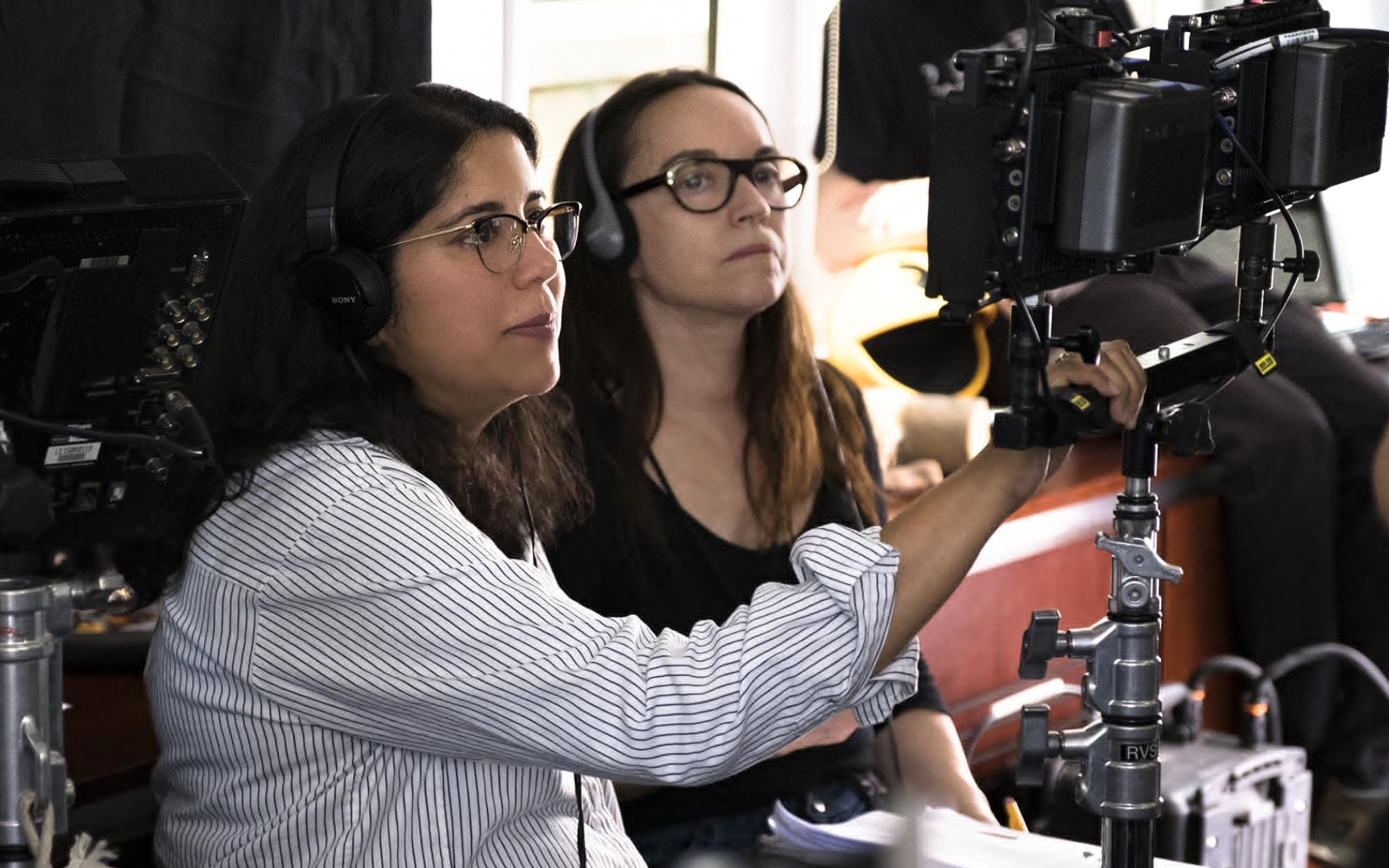 Two women wearing headsets are focused on a film camera setup, working behind the scenes on a production set