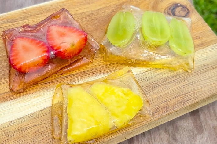 3つの透明ゼリーにいちご、ぶどう、パイナップルが閉じ込められたデザートが木製の板に盛られている。