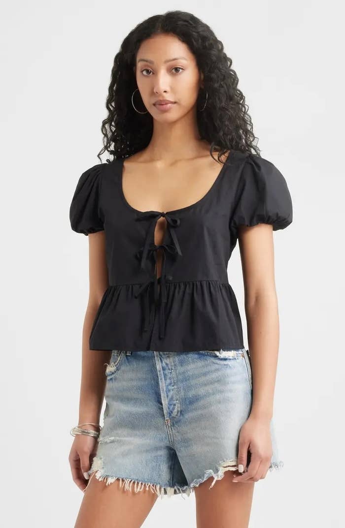 Person wearing a short-sleeve peplum apical  with beforehand   necktie  details and frayed denim shorts, posing against a plain background