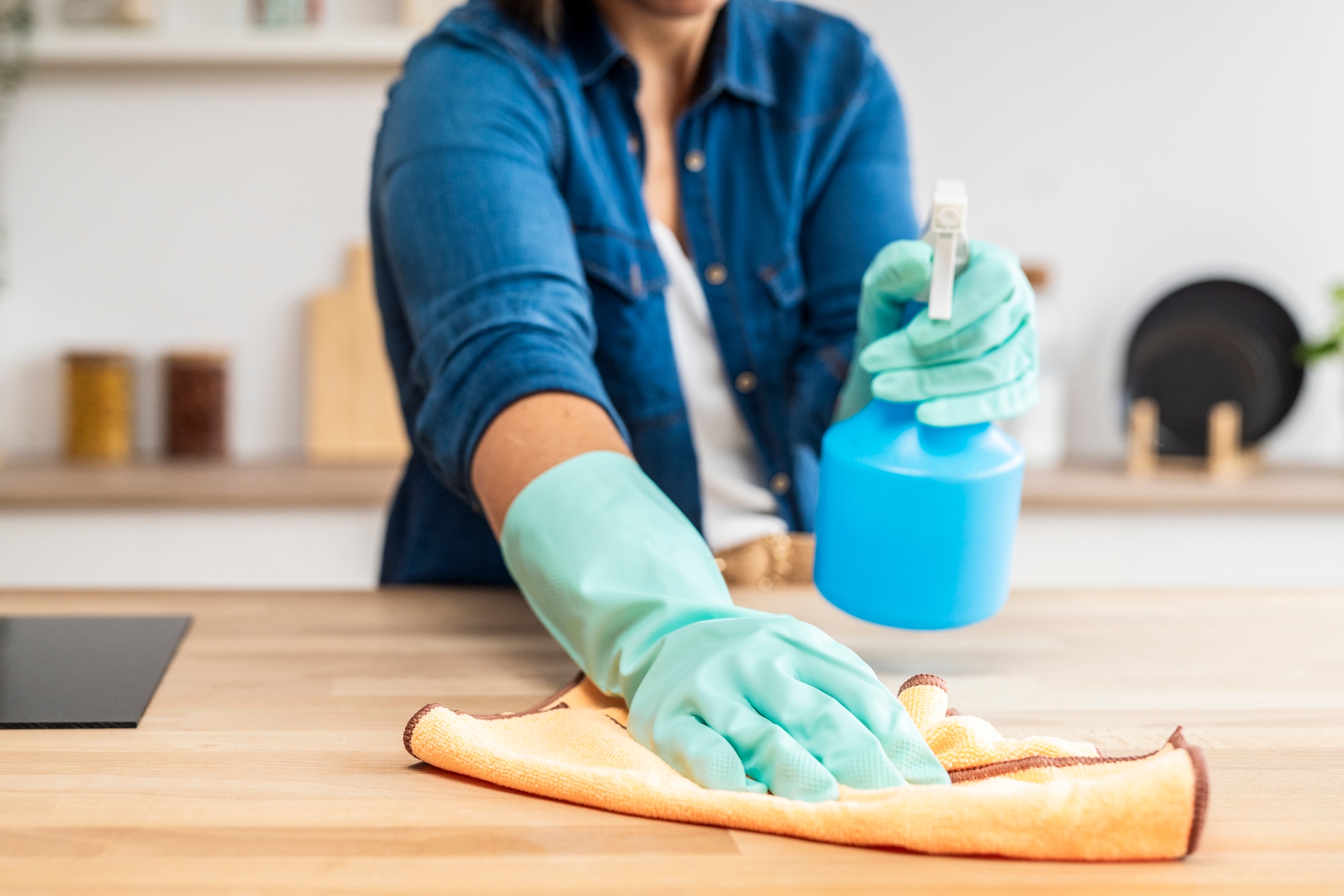 A person cleaning a kitchen countertop with a spray bottle and cloth, wearing gloves for protection and hygiene