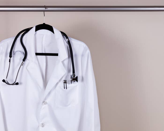 A white lab coat with a stethoscope draped over the shoulders hangs on a hanger against a plain background