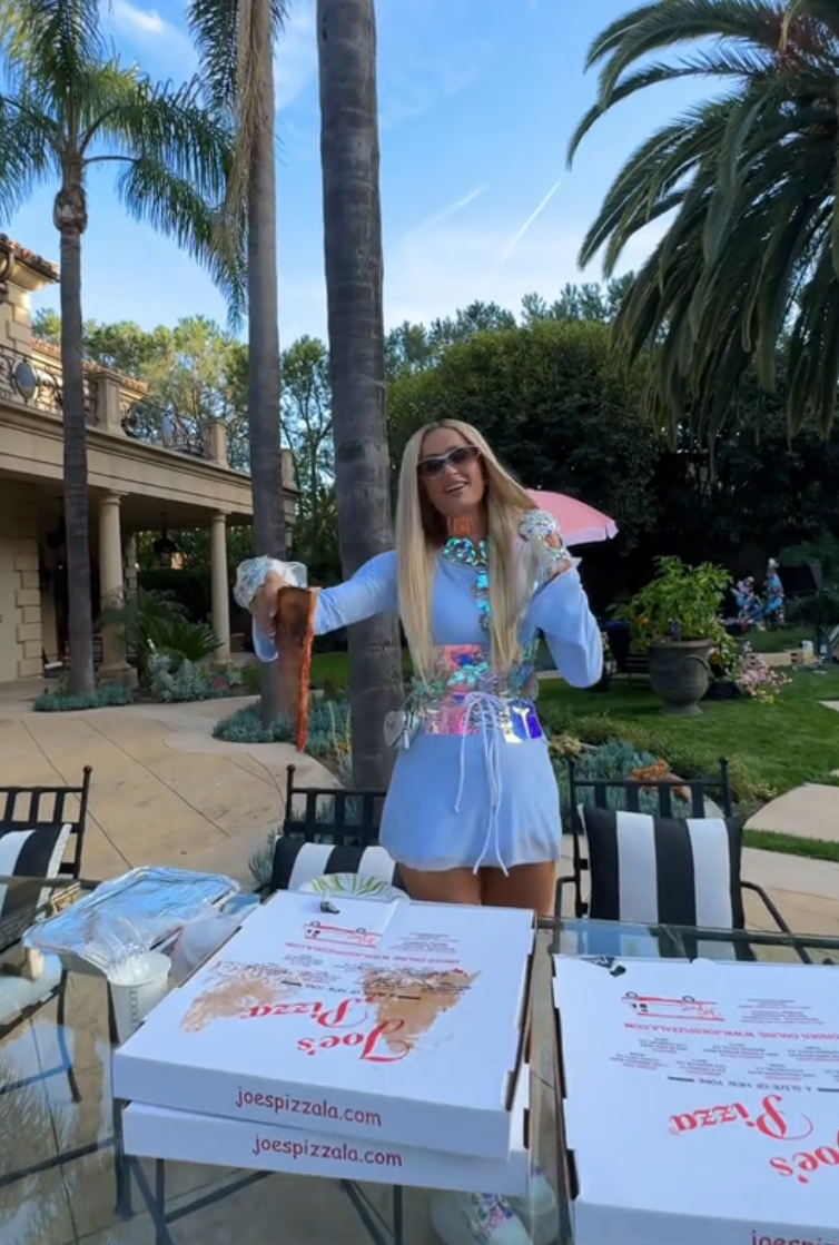 Paris Hilton holding a pizza slice above pizza boxes outdoors