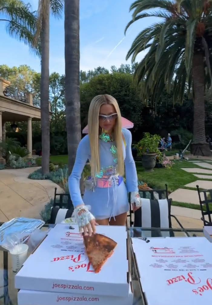 Person with long hair holds a slice over pizza boxes outdoors, surrounded by palm trees. They wear a stylish, casual outfit