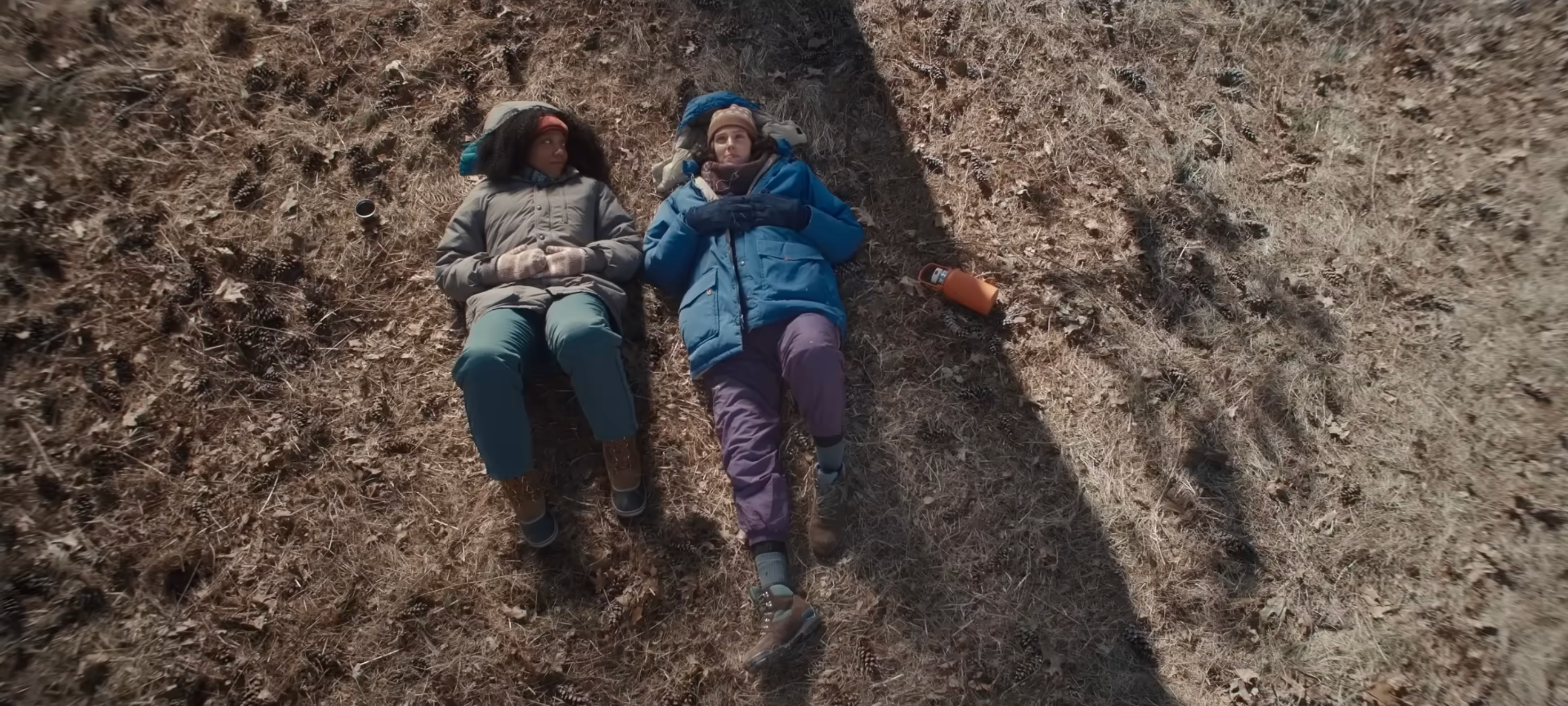 Two people in winter clothing lie on dry grass, looking upwards. A thermos is nearby