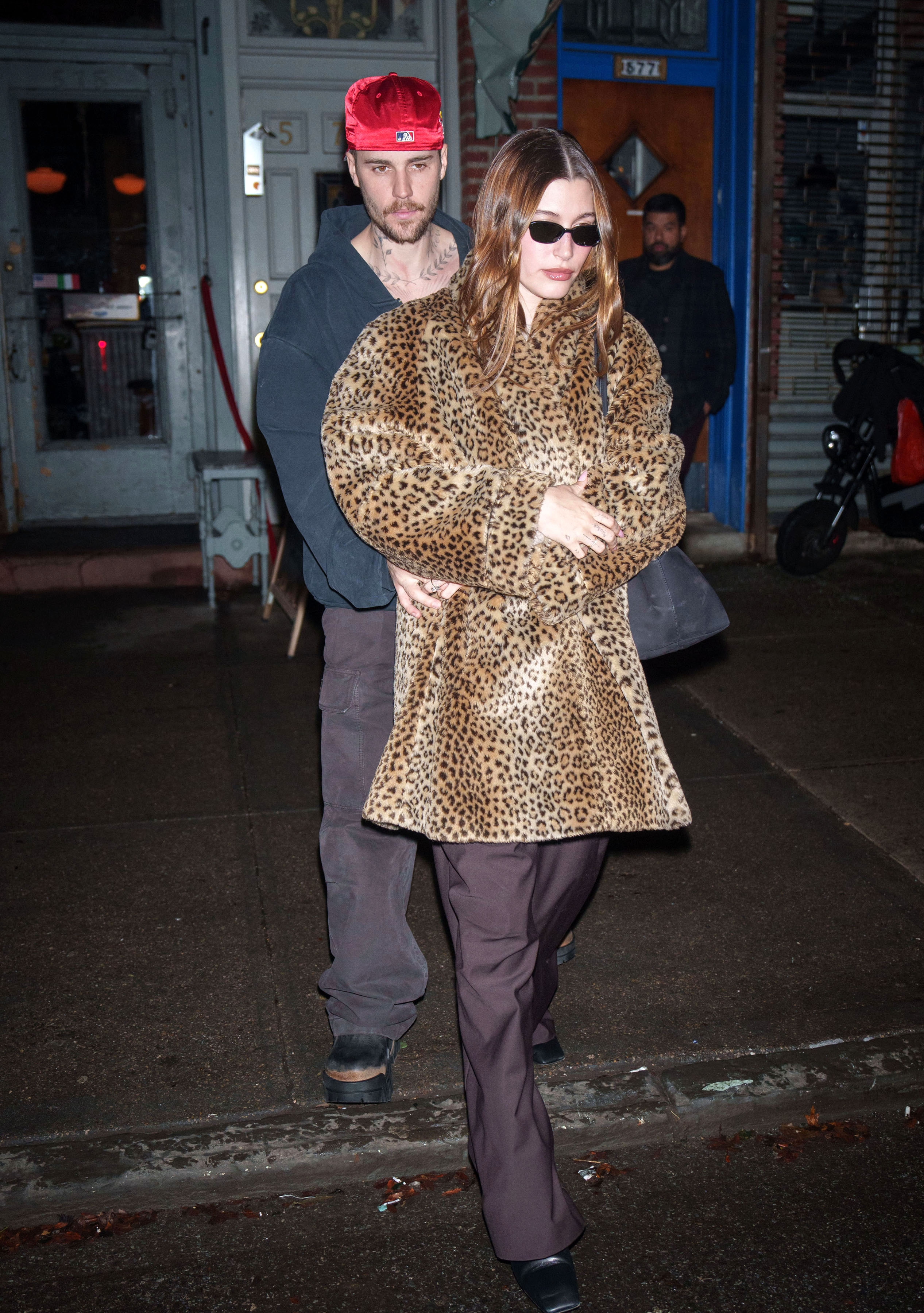 Person in a leopard print coat and sunglasses walks on a sidewalk, followed by another person in a casual outfit and red cap