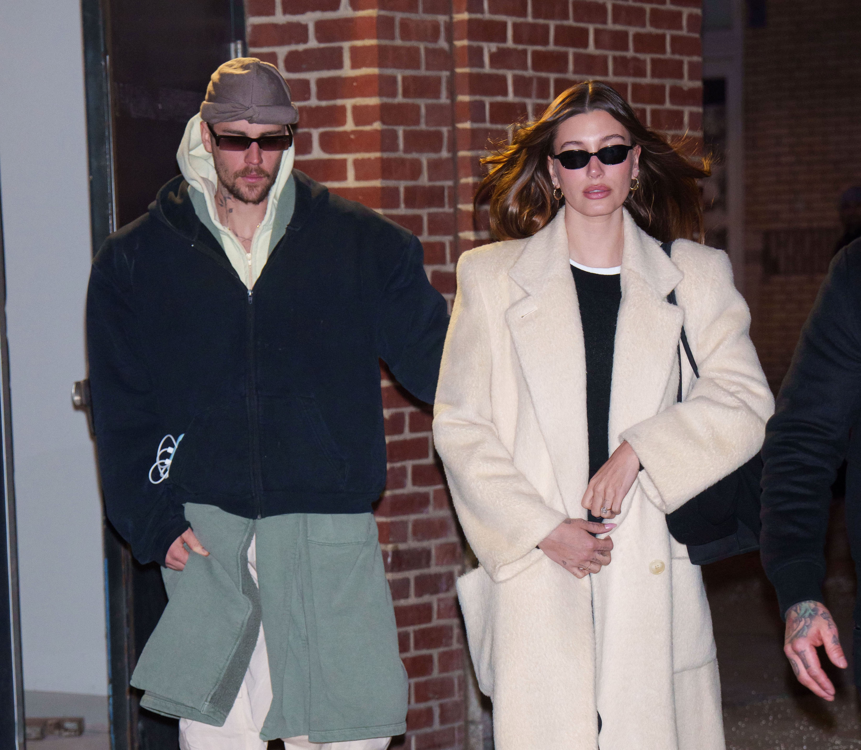 Two individuals in stylish, casual attire wearing sunglasses. The person on the left wears a hoodie and pants, while the person on the right wears a long coat