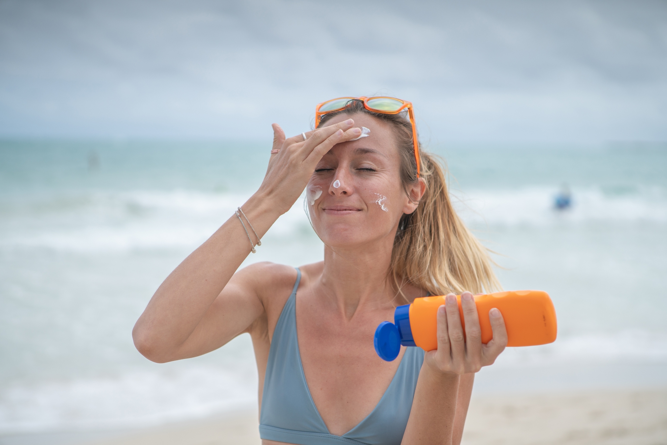Person at the beach applying sunscreen on their face, smiling, wearing a bikini and holding a sunscreen bottle