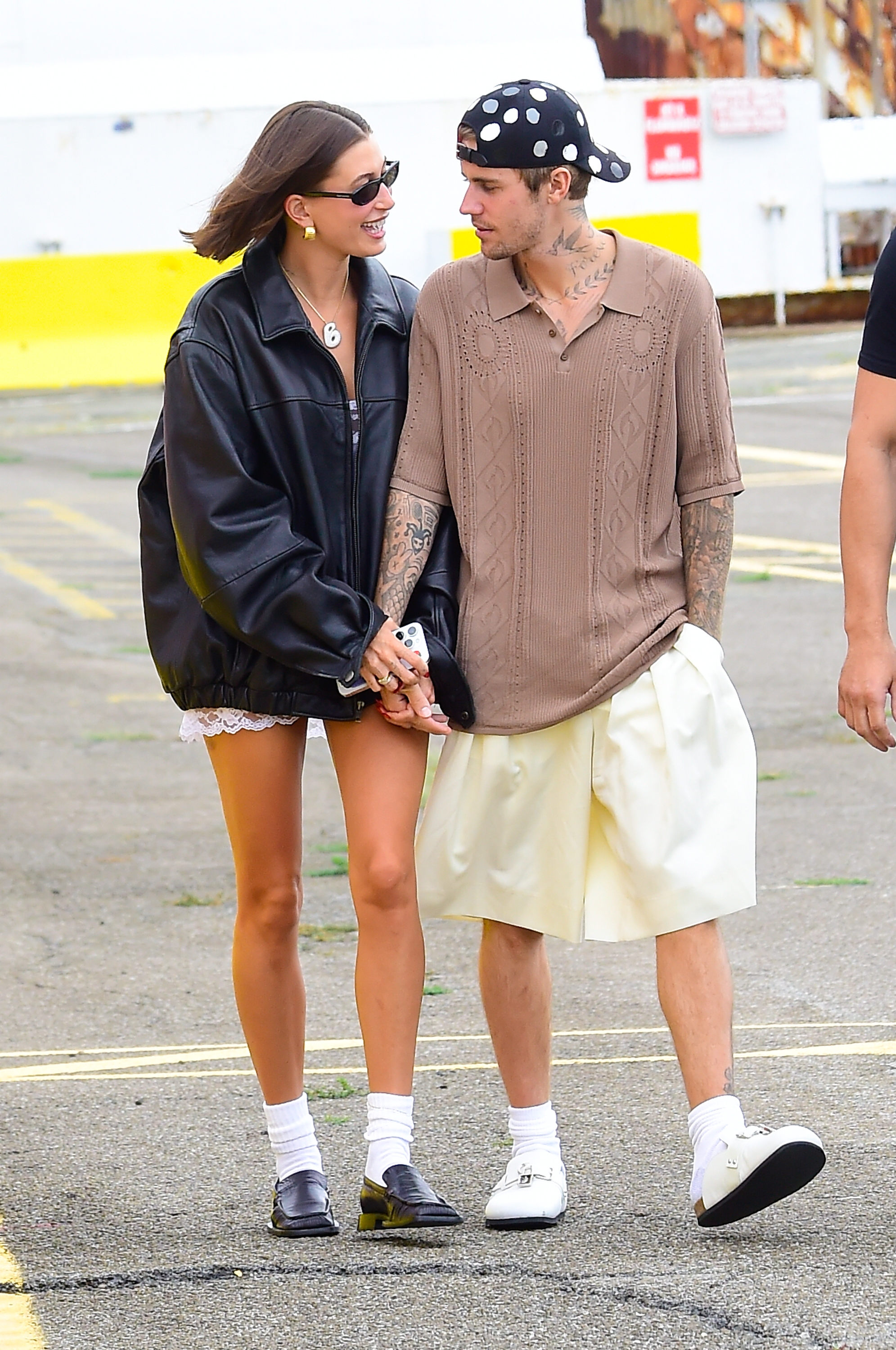Two individuals holding hands, wearing casual streetwear: oversized leather jacket with shorts, and knit polo with baggy shorts, tattooed arms visible