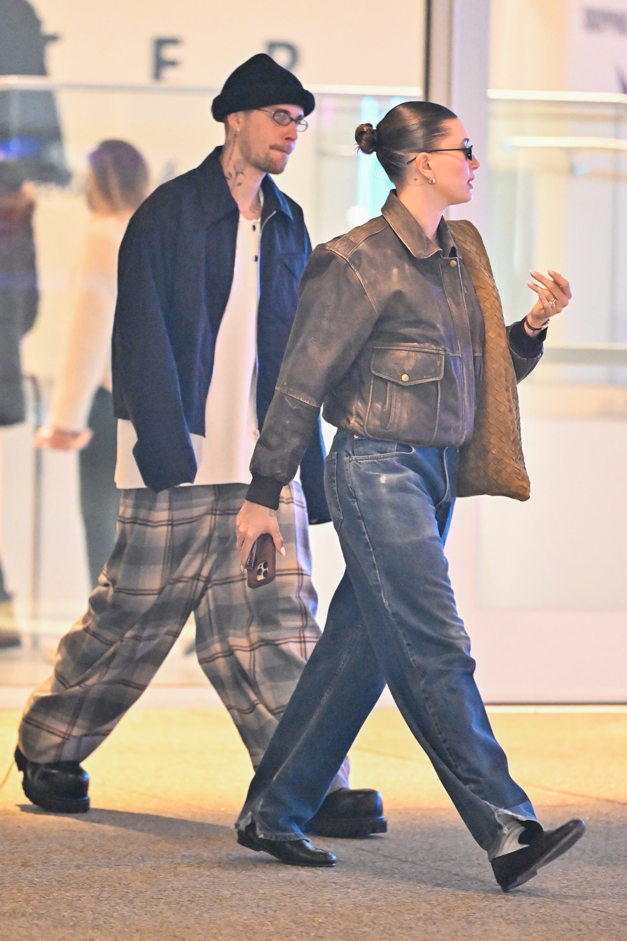 Two individuals walk indoors. One wears a beanie, plaid pants, and a dark jacket. The other wears sunglasses, a leather jacket, and holds a bag