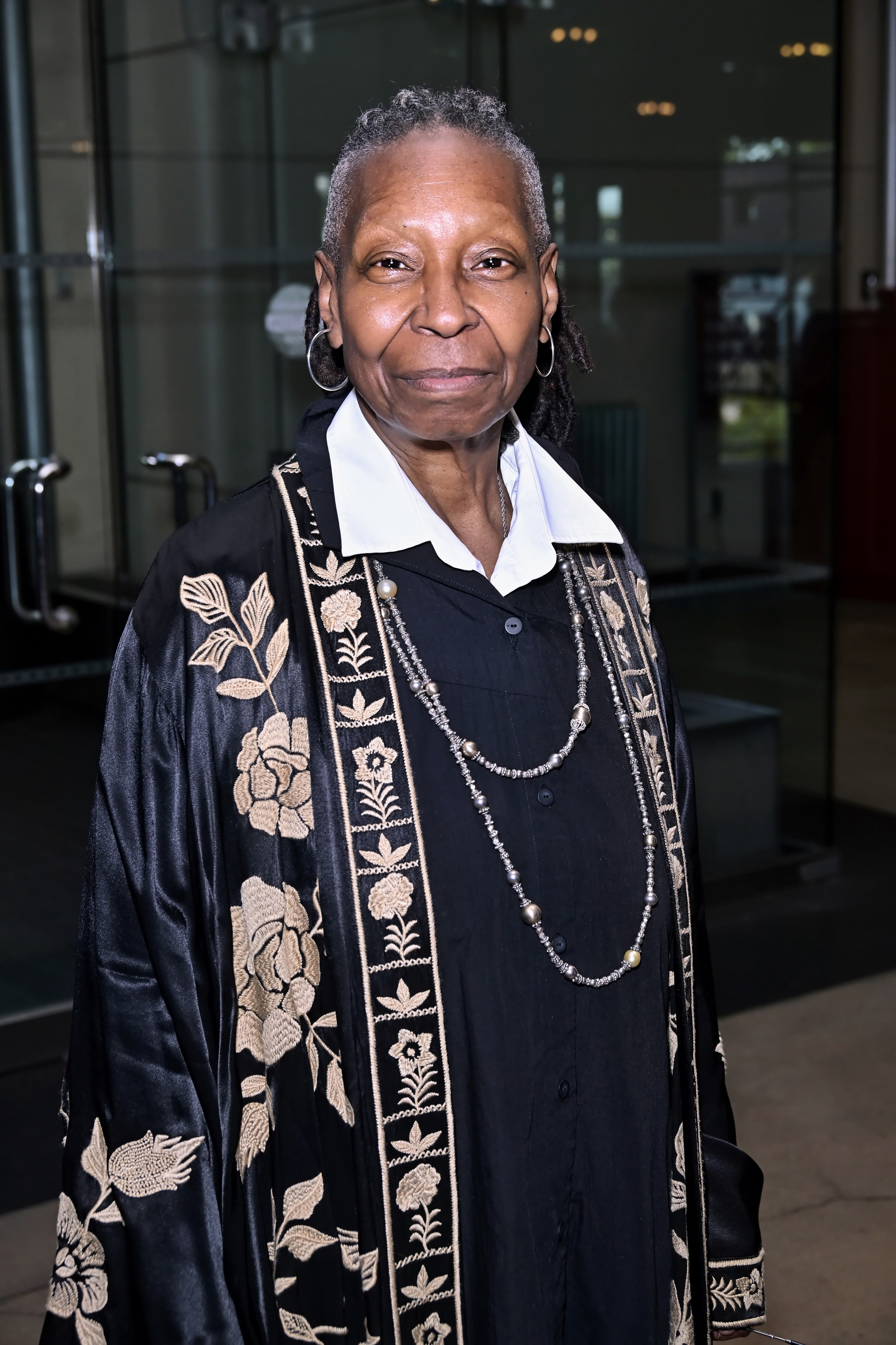 Celebrity in an embroidered coat over a collared shirt, standing indoors, looking at the camera