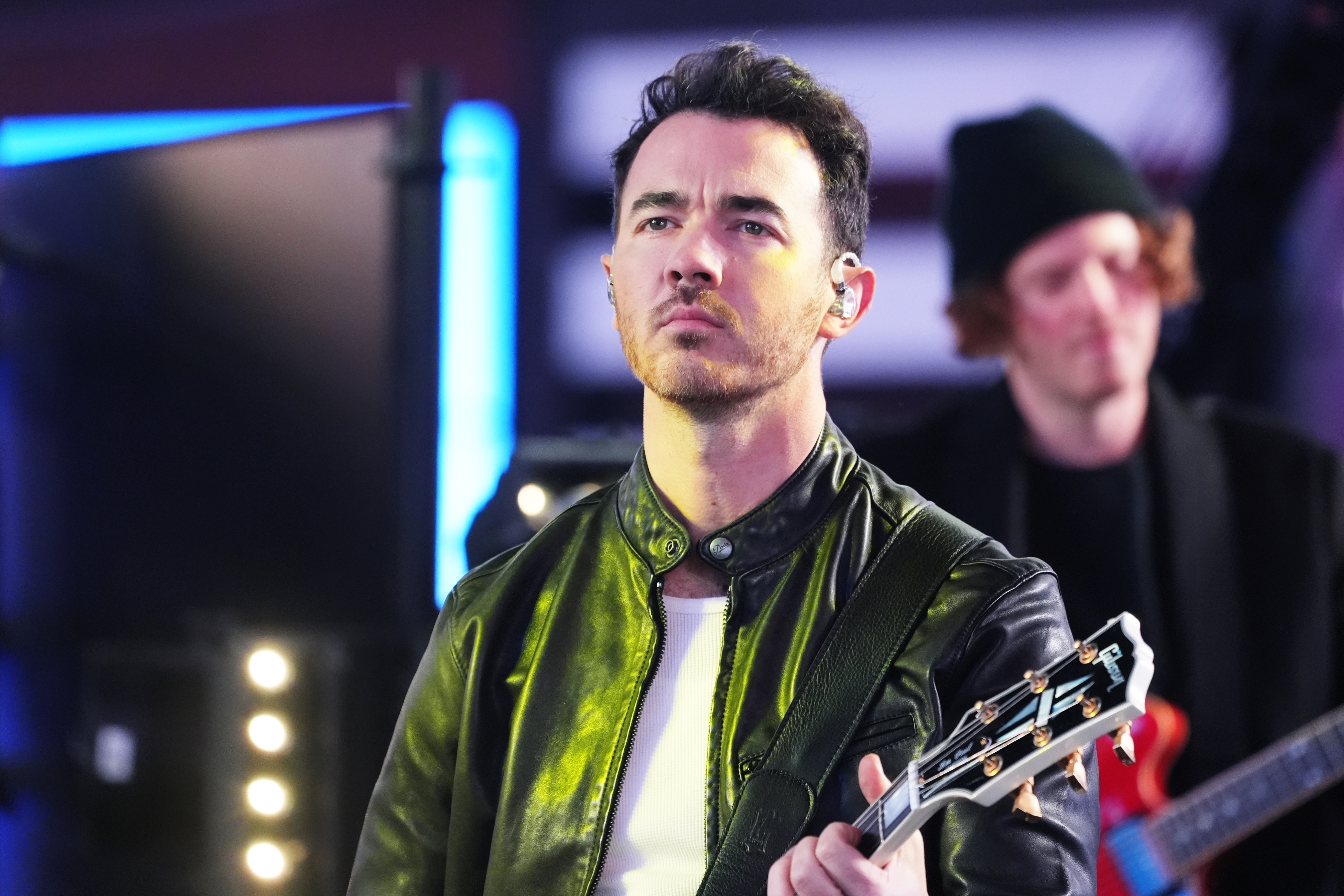 Musician holding a guitar performs on stage, wearing a leather jacket. Another band member with a beanie is in the background