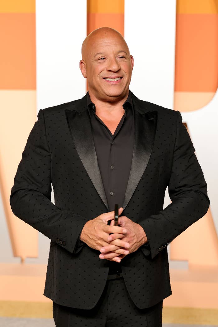 Person in a textured black suit with shiny lapels, smiling on a red carpet