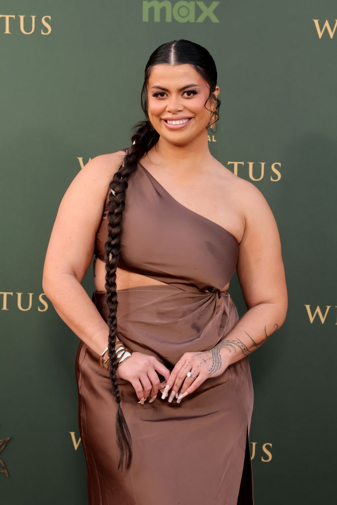 Person on a red carpet wearing an elegant, asymmetrical gown with a long braid and tattoos on one arm