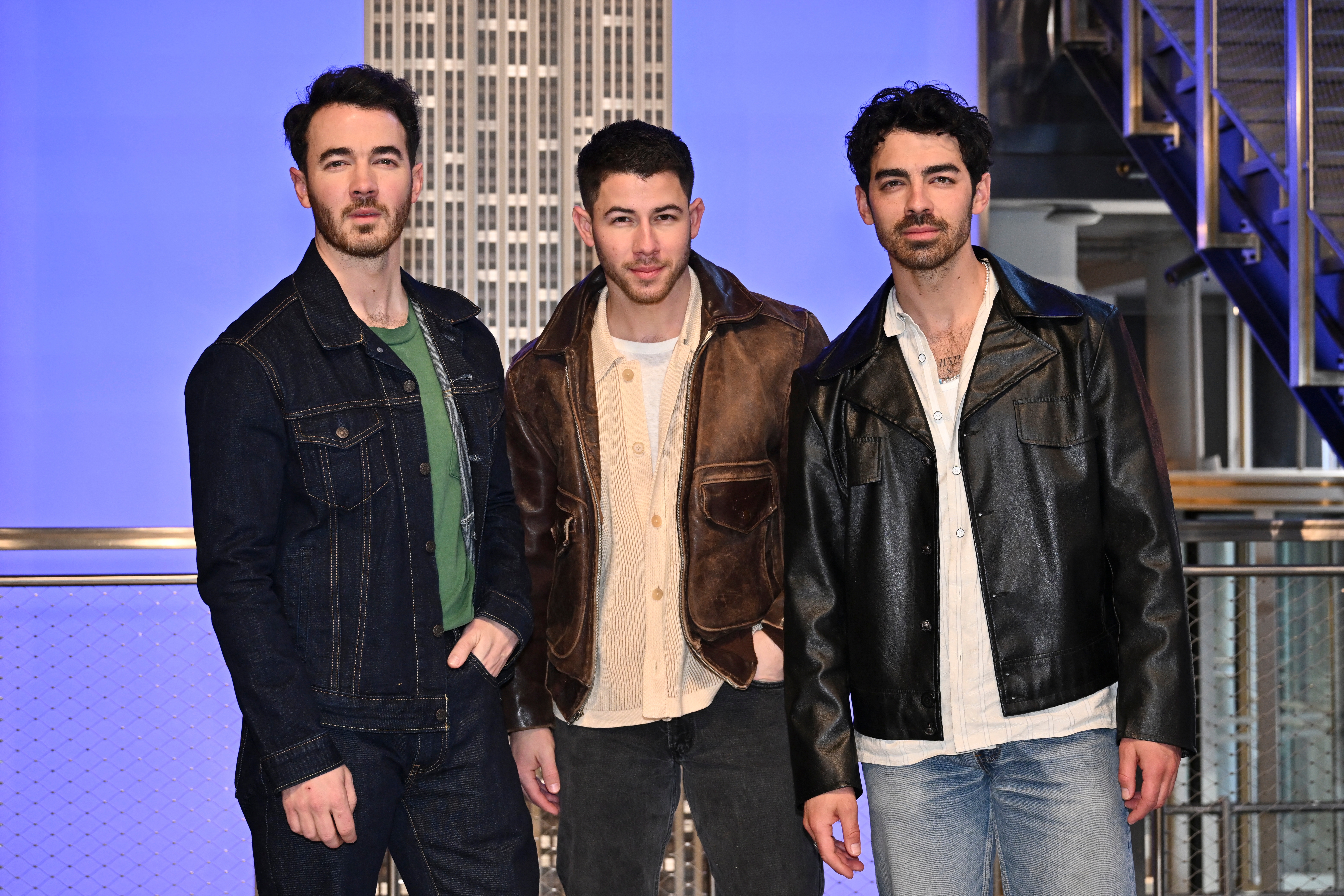 Three men in stylish jackets and casual shirts pose indoors with a cityscape backdrop