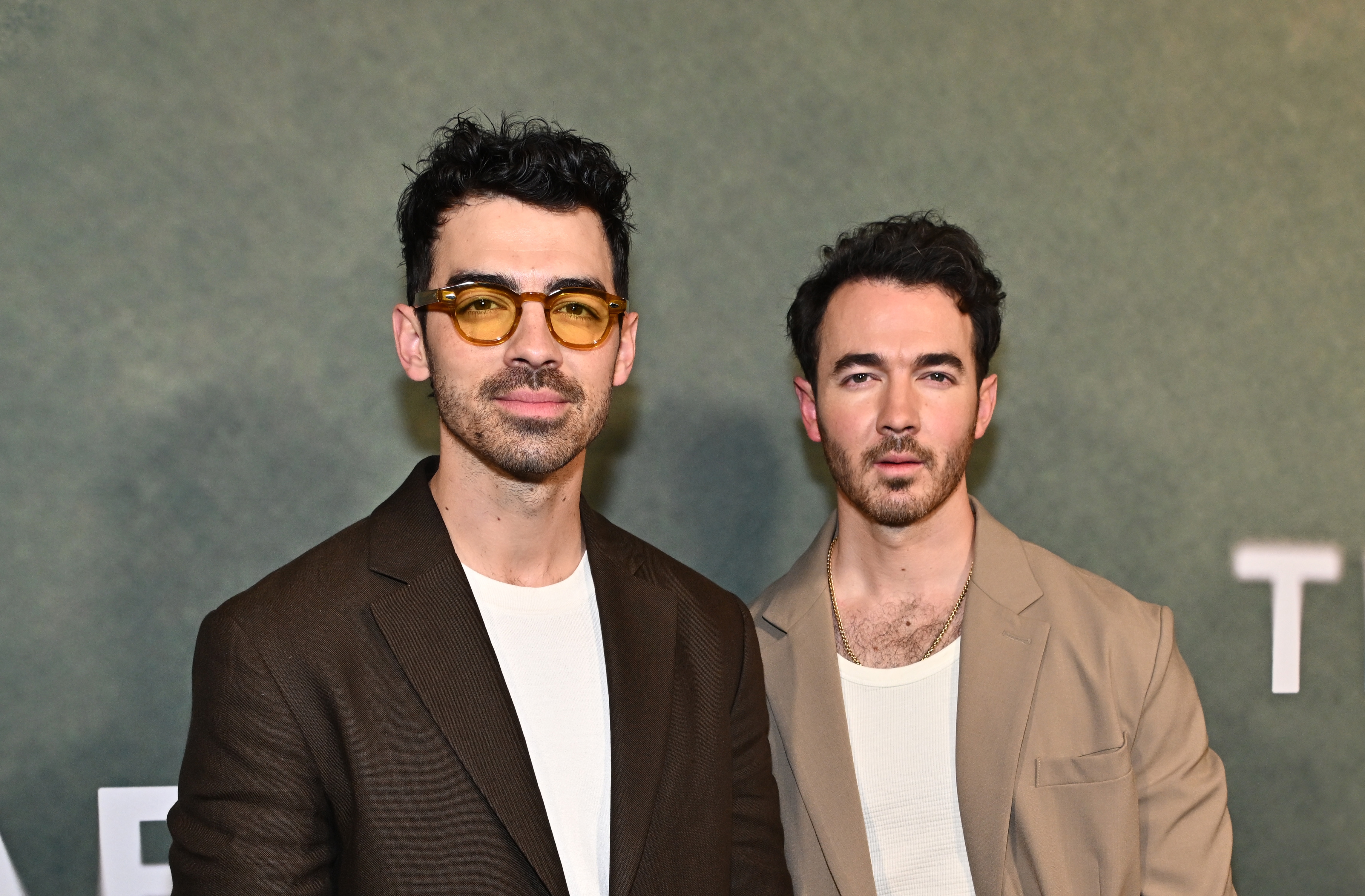 Two individuals at an event; one wears a brown jacket and yellow-tinted glasses, the other a beige jacket, both with white shirts