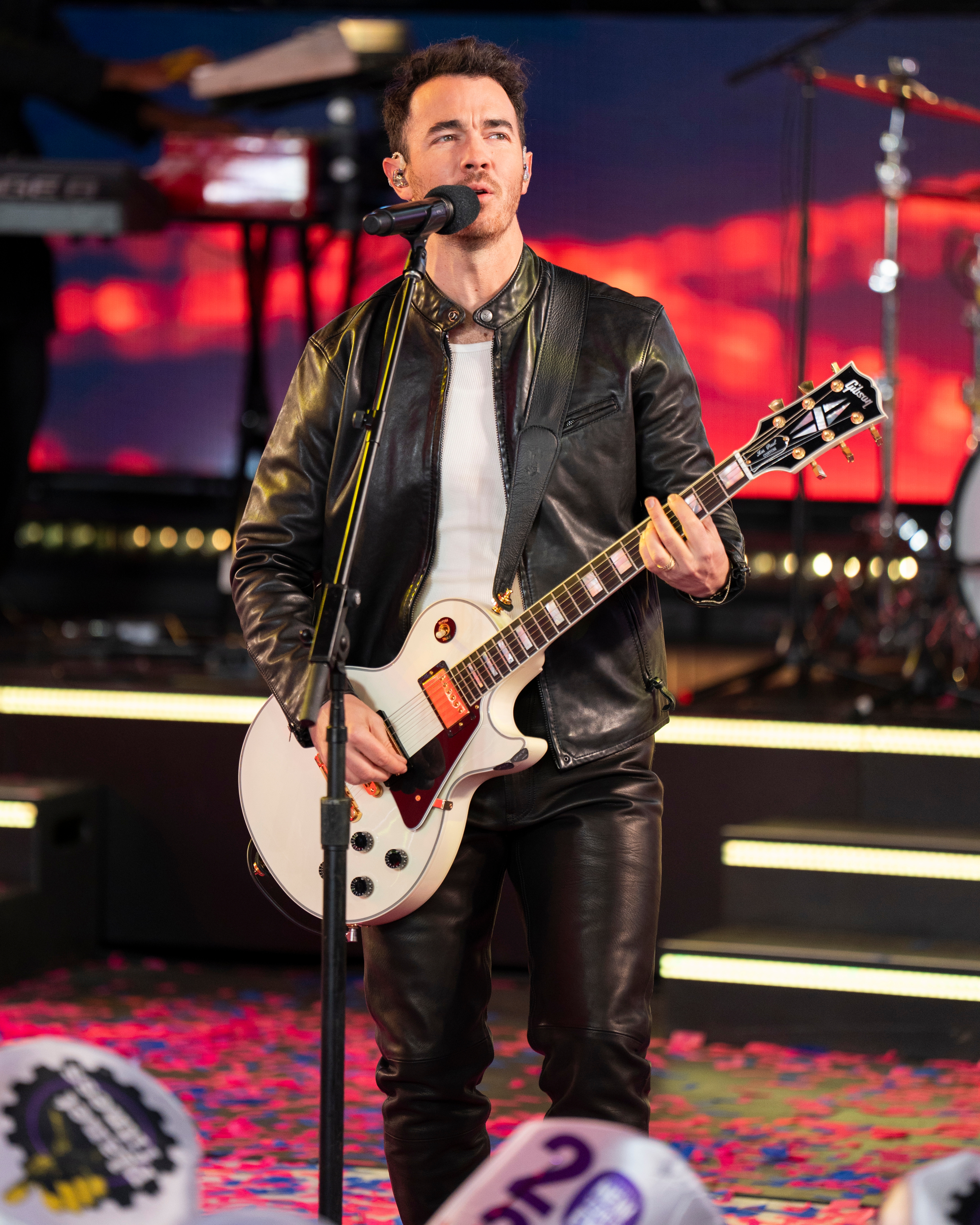 Musician performs on stage with electric guitar, wearing a leather jacket and pants, under colorful stage lights