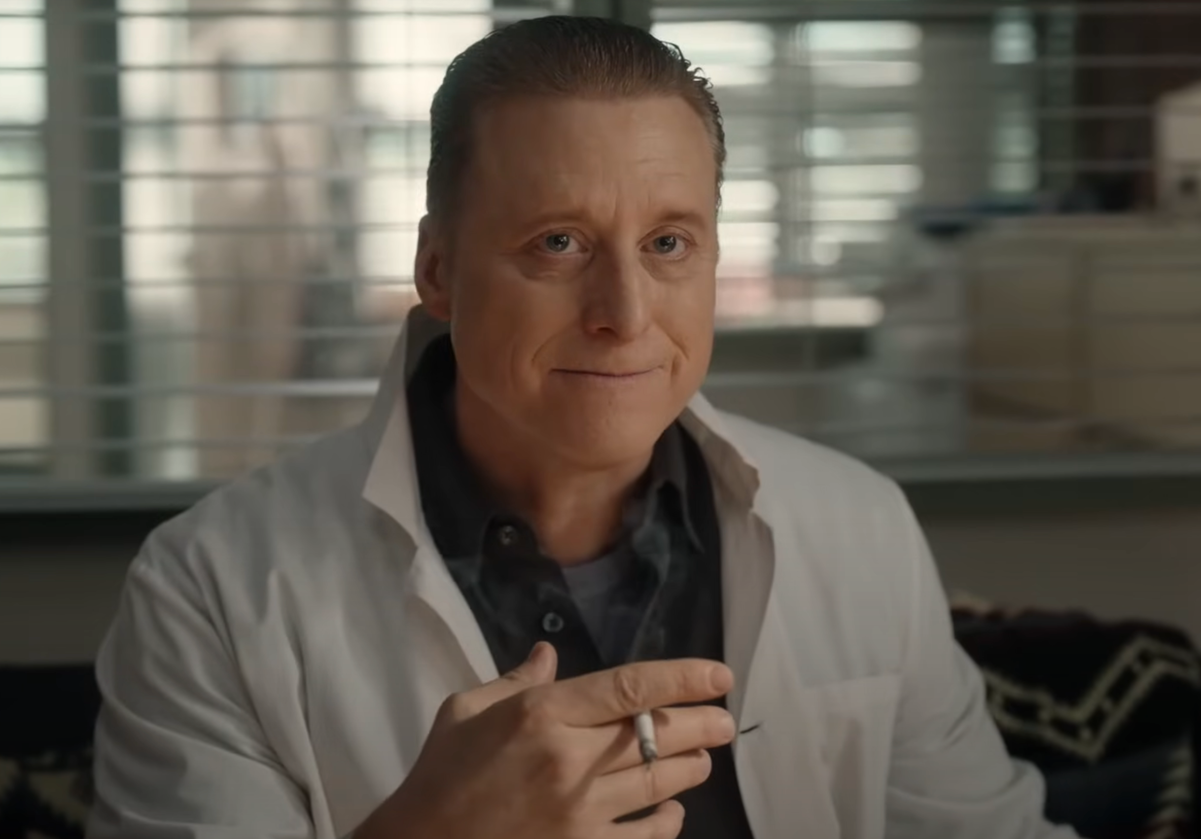 Person in a lab coat holds a pen, seated indoors with a window in the background