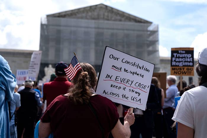 Protesters gather outside a government building; a sign reads, "Our Constitution must be upheld in every case; keep it strong across the USA."