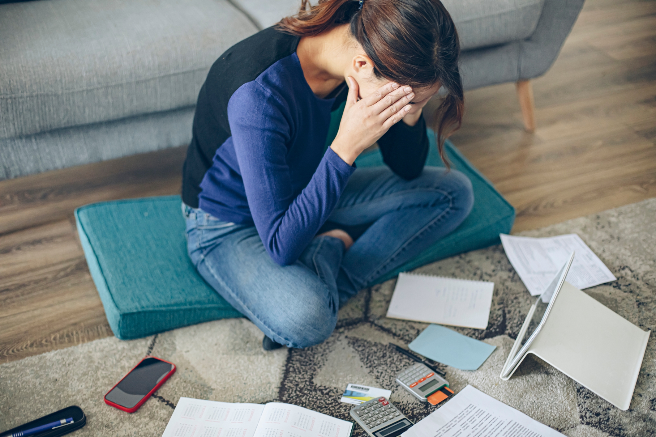 Person sitting on the floor, holding head in hands, surrounded by scattered bills, a laptop, and a phone, appearing stressed or overwhelmed