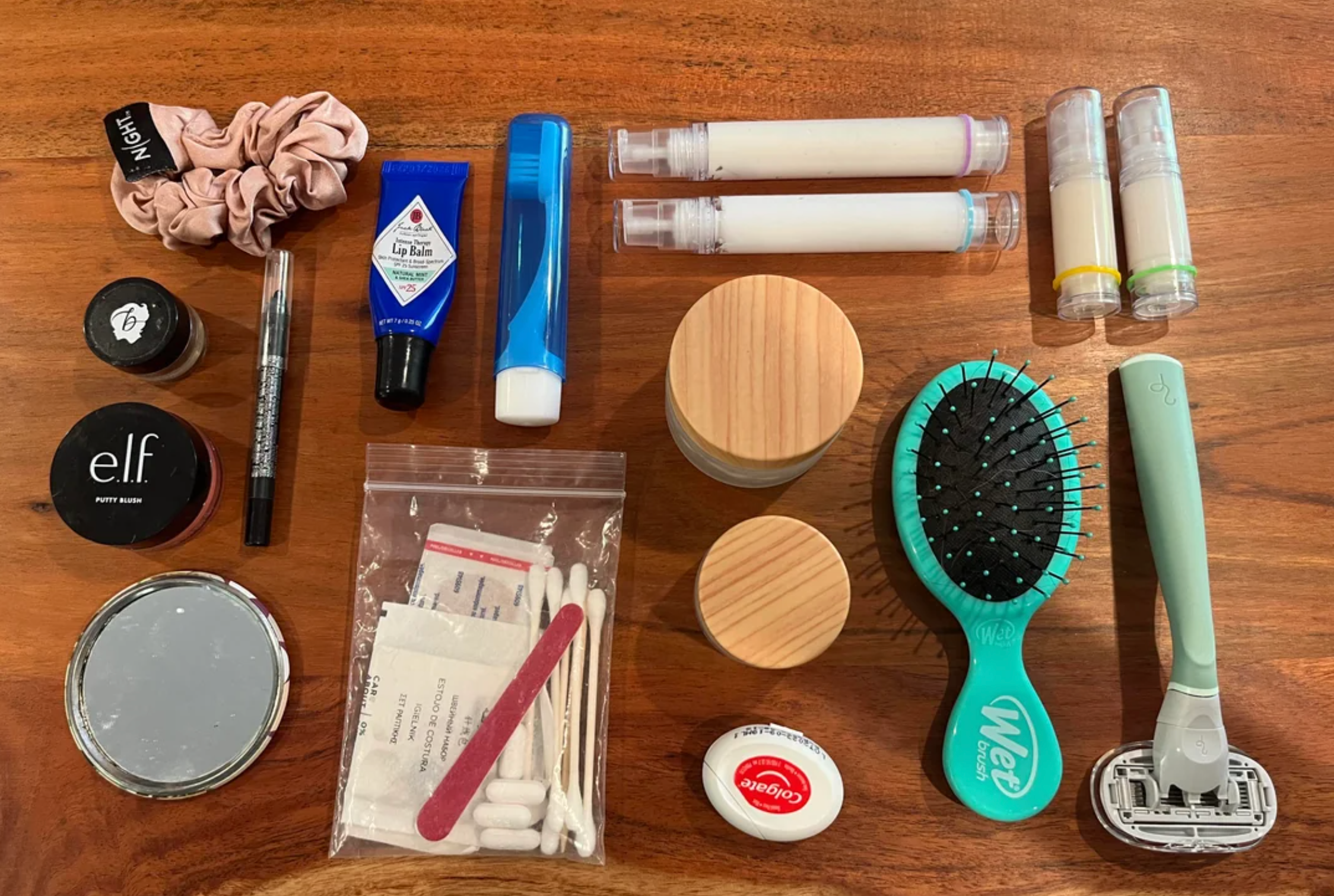 Collection of toiletries and personal care items on a wooden surface, including makeup, hairbrush, lip balm, sunscreen, and hygiene products