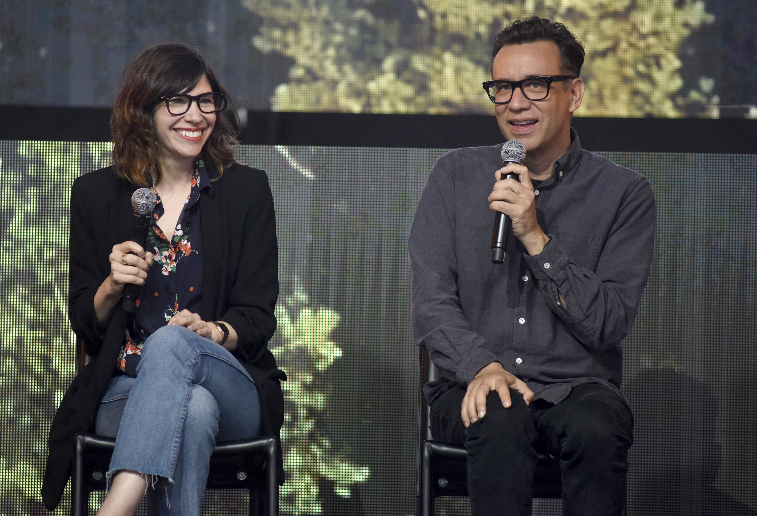 Fred Armisen and Carrie Brownstein sitting on stage, holding microphones, one wearing a floral blouse and blazer, the other in a casual shirt. Smiling and engaging audience