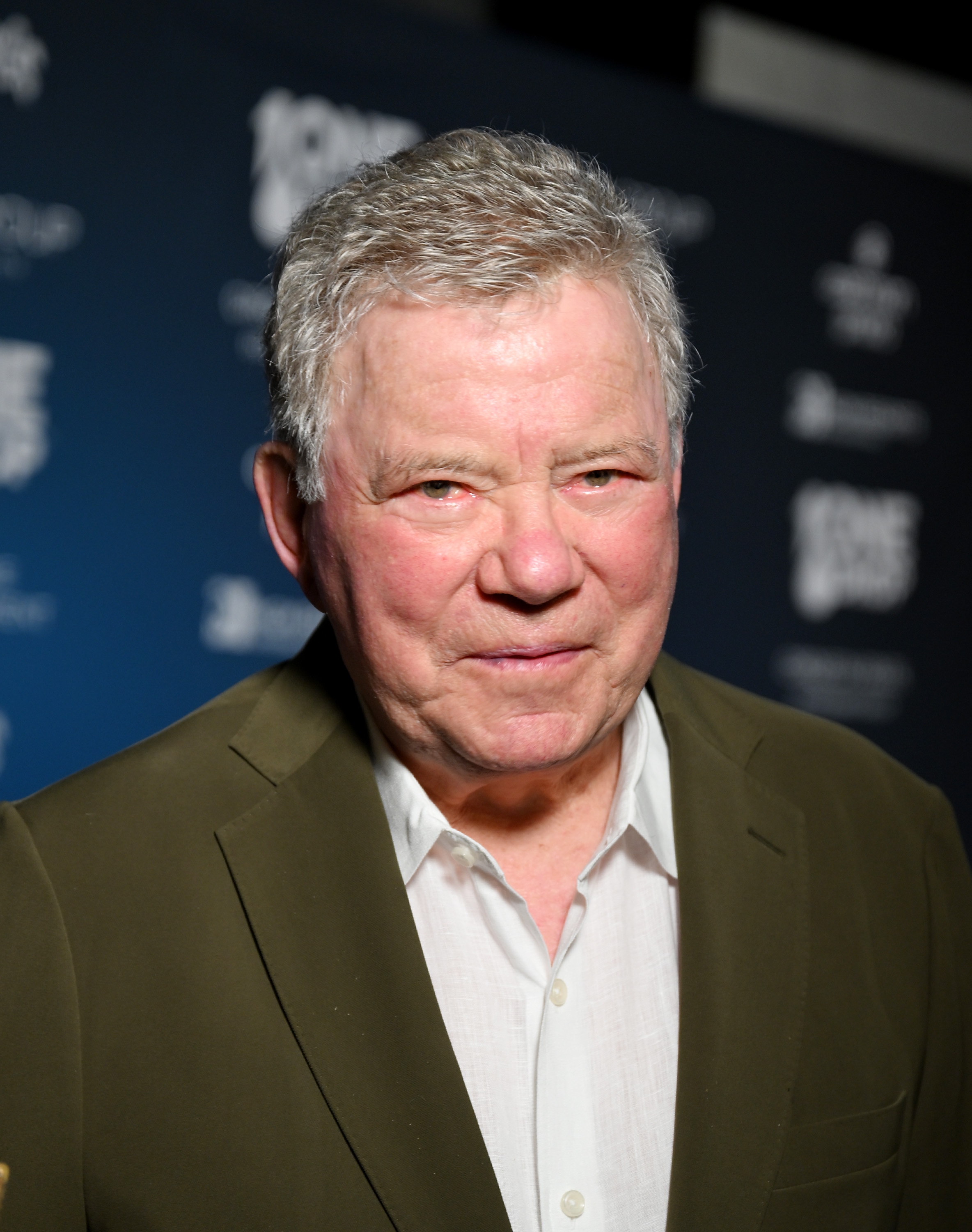William Shatner in a white shirt and jacket poses on an event red carpet