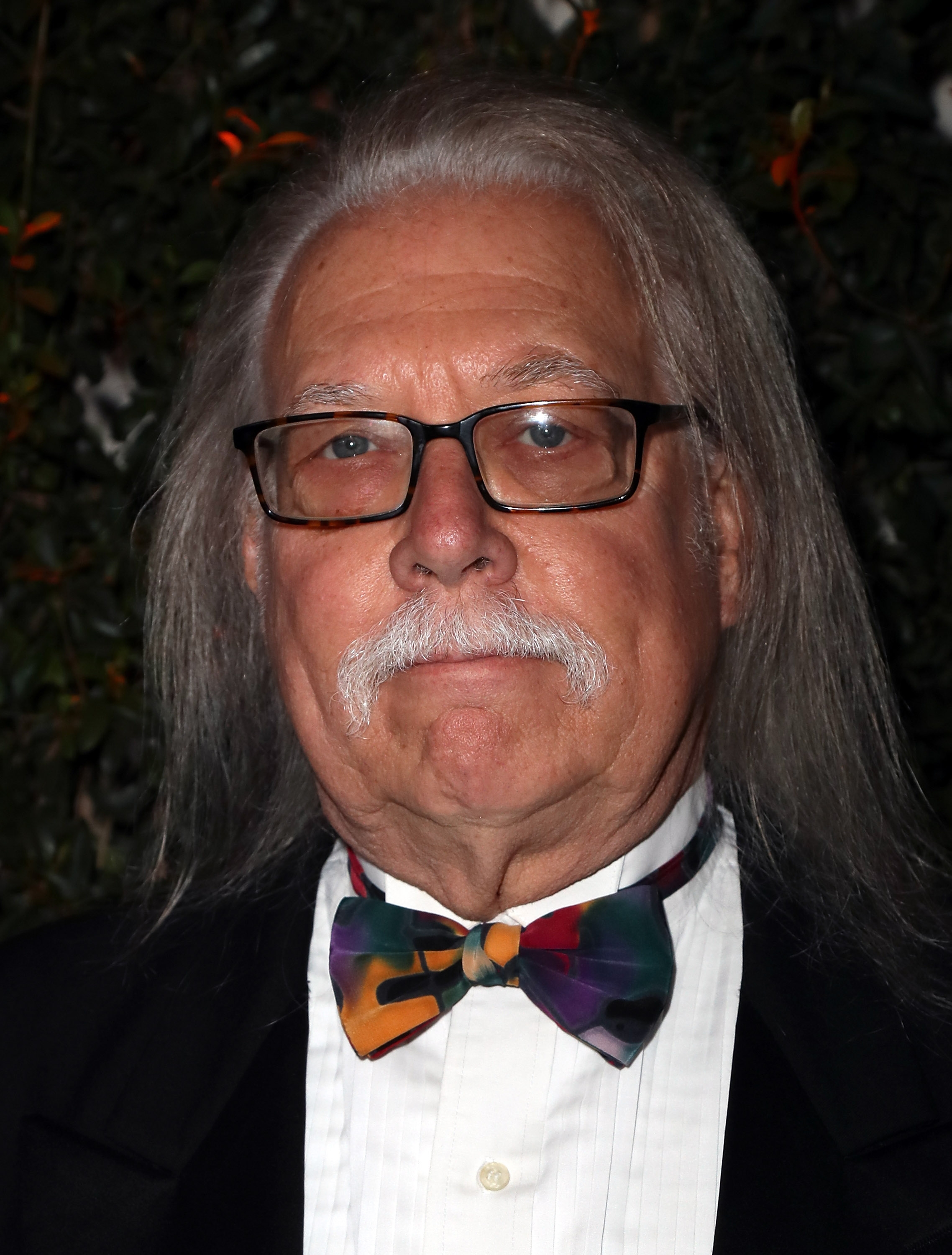Bob Zmuda with long hair, wearing glasses, a tuxedo, and a colorful bow tie at a formal event