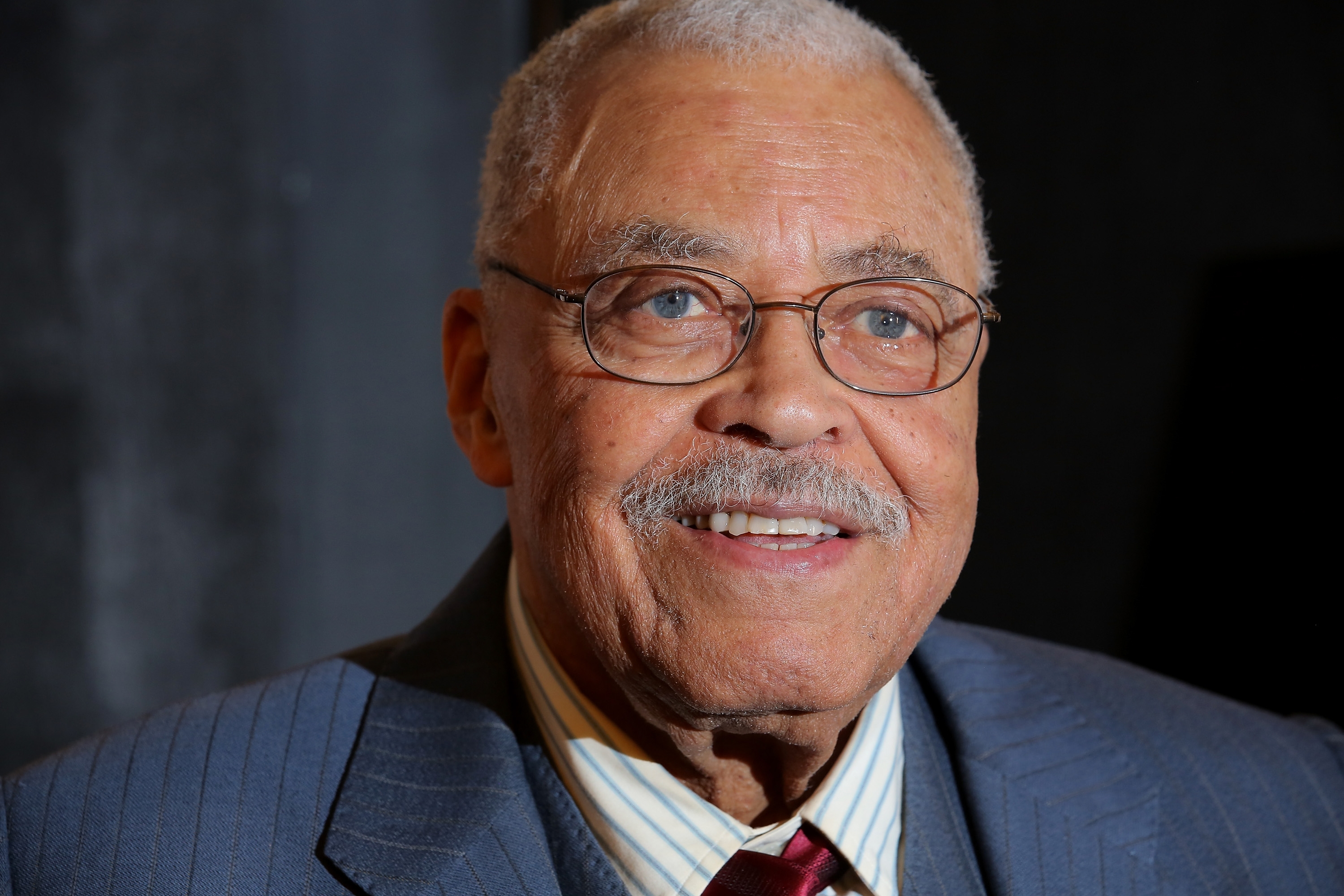 Elderly man in glasses, with a mustache, wearing a striped shirt, a tie, and a suit jacket, smiling at a formal event