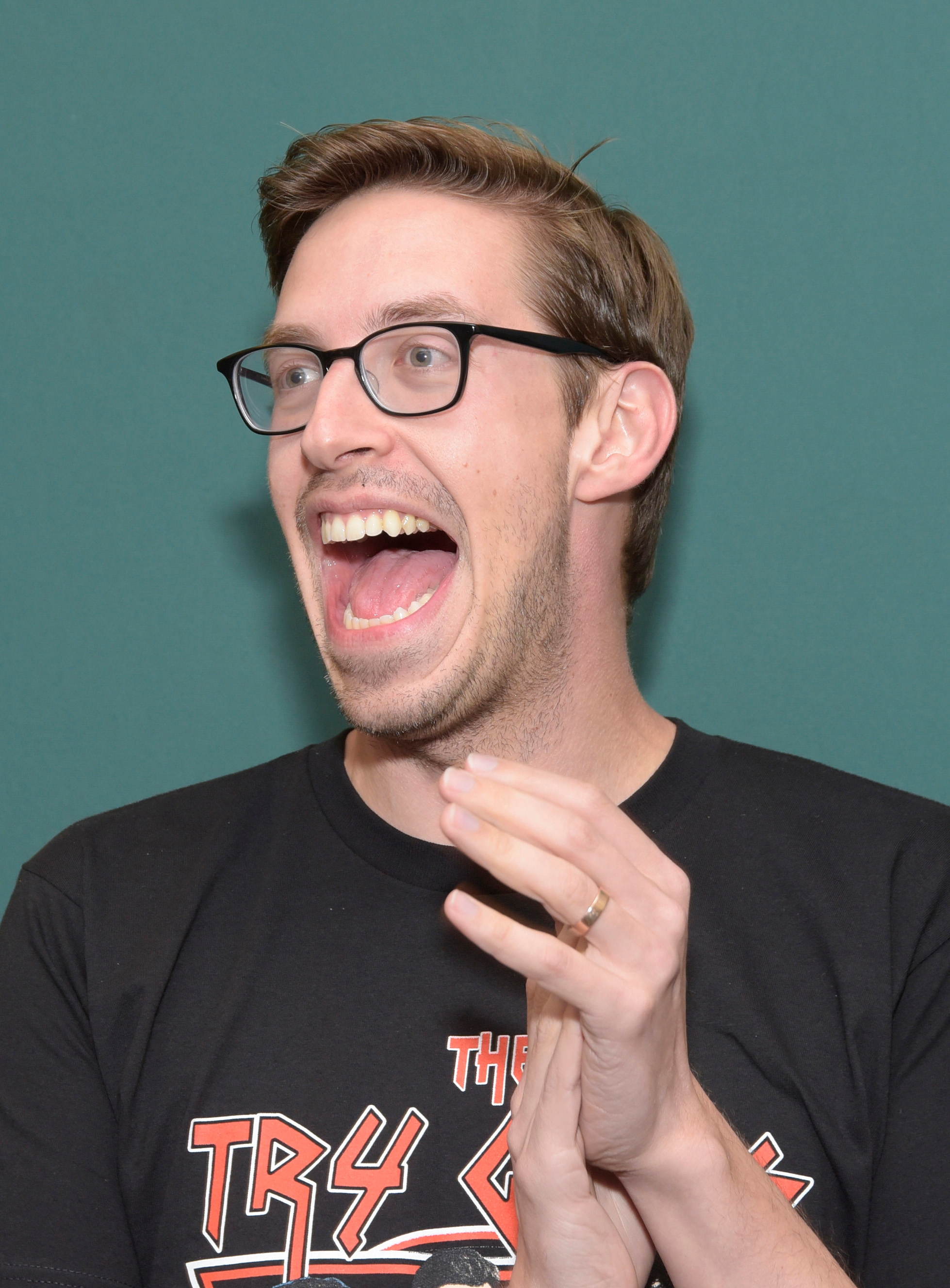A person wearing glasses and a graphic T-shirt is laughing with hands raised in a lively gesture