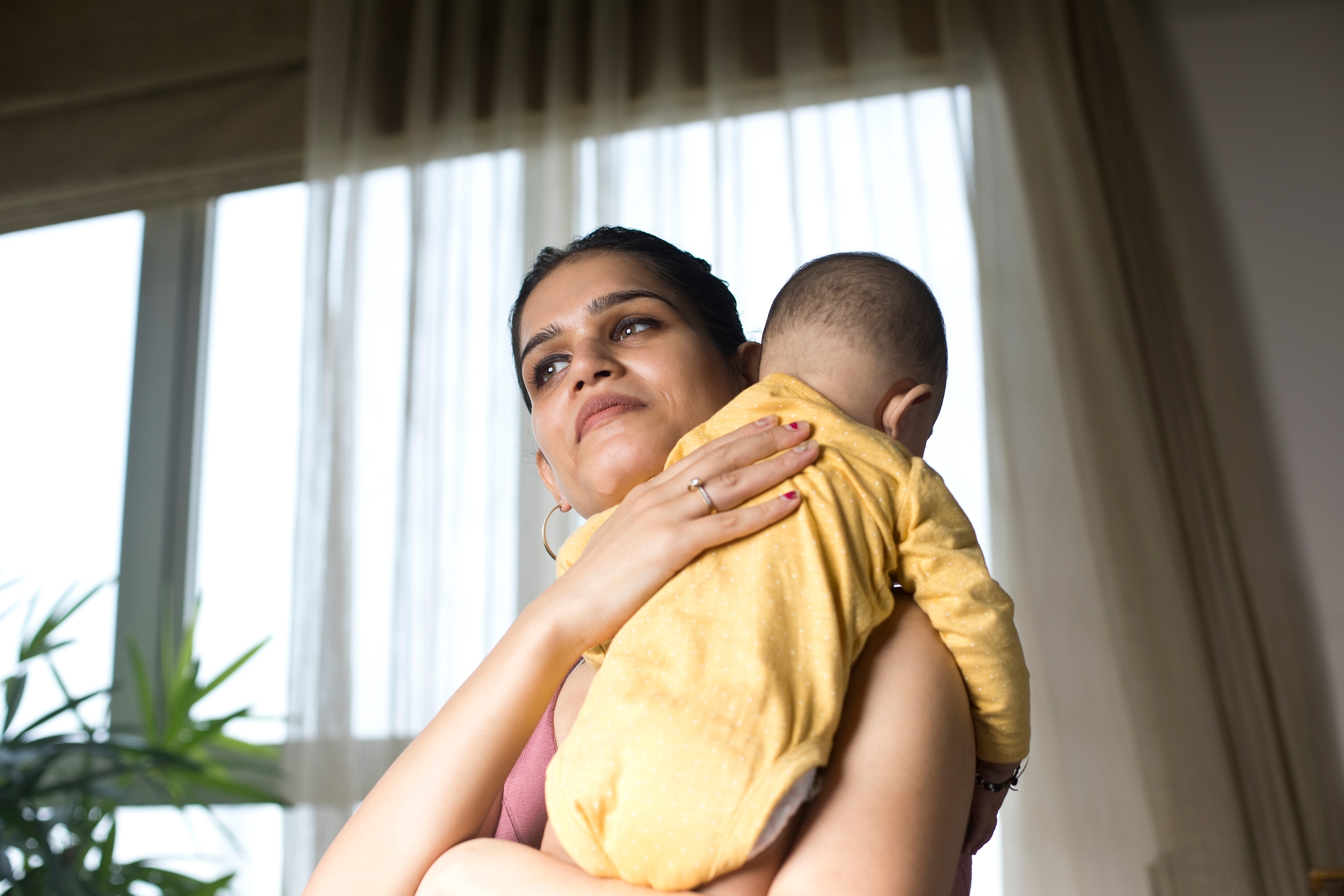 A person gently holds a baby wearing a onesie, standing by a window with curtains, conveying a tender moment