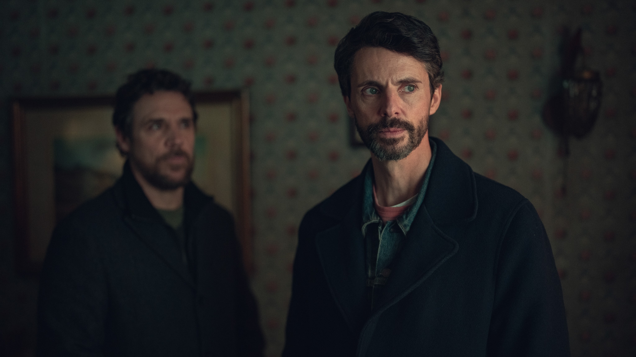 Two men in a dimly lit room appear concerned. Both wear dark coats, one with a beard, suggesting a tense movie or TV scene