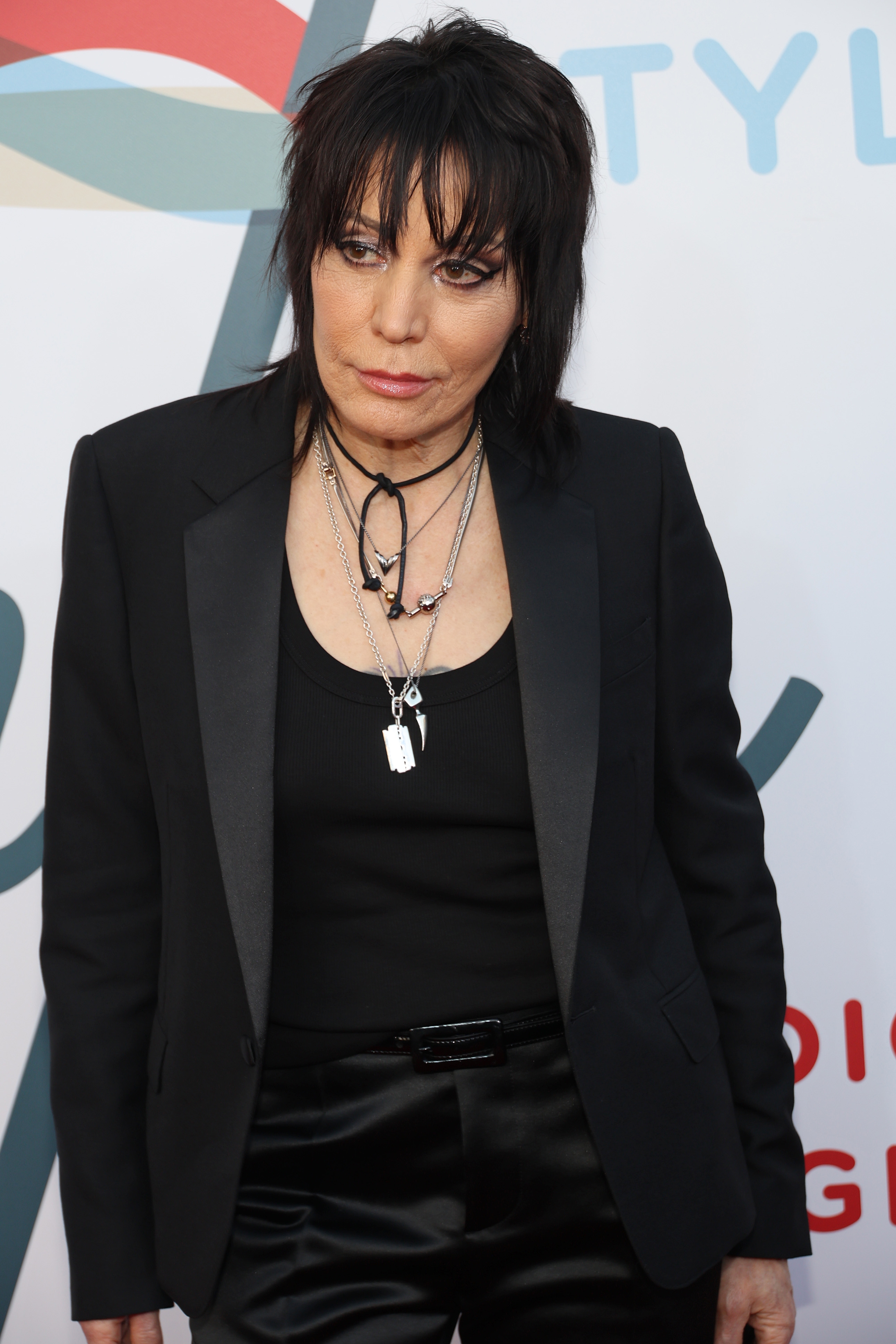 Joan Jett in a black suit with layered necklaces at a formal event, standing against a backdrop with stylized writing