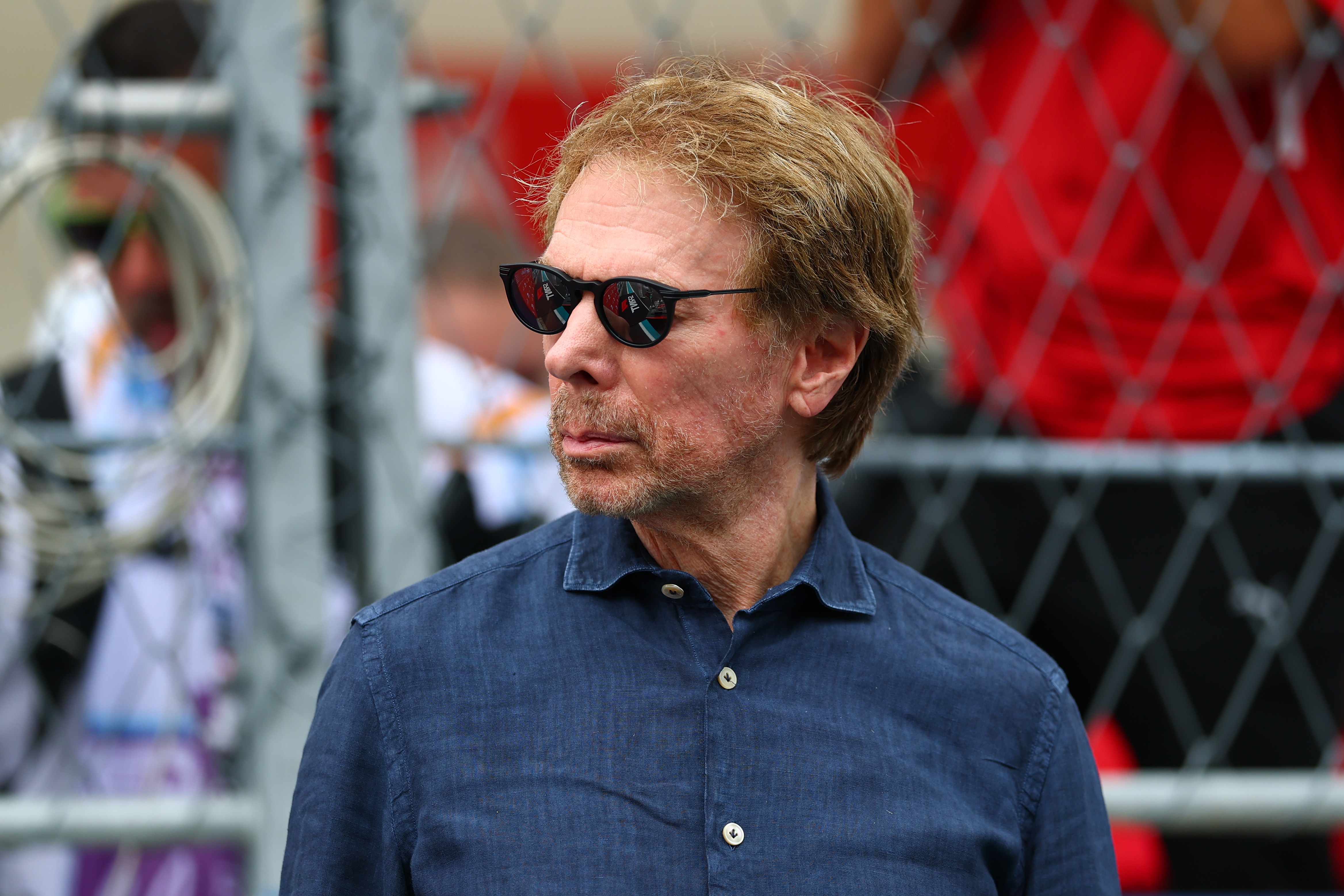Jerry Bruckheimer wearing sunglasses and a dark shirt stands in front of a chain-link fence, looking to the side. The background includes blurred figures