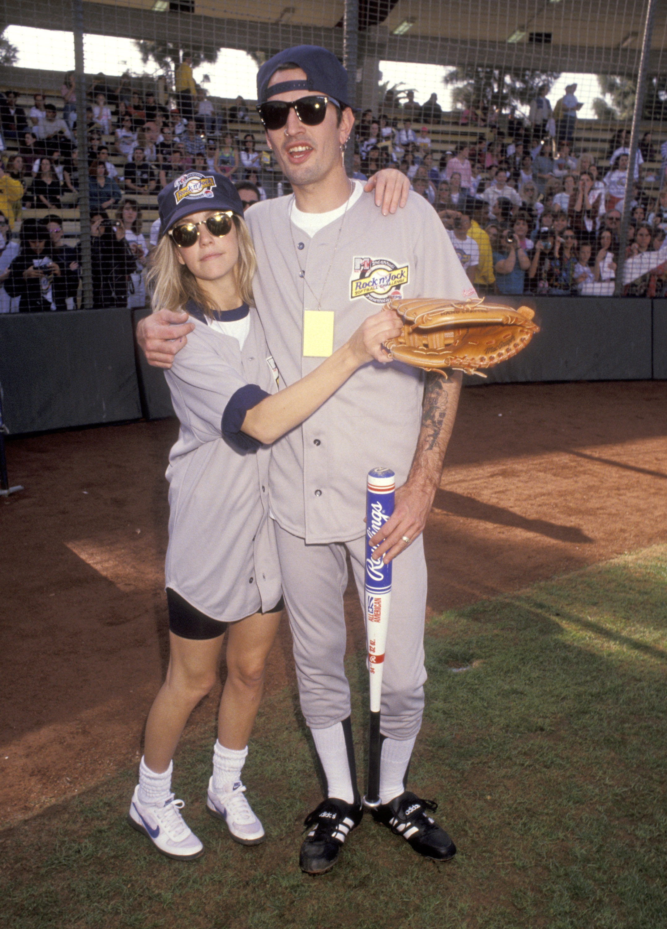 Tommy Lee, Heather Locklear in baseball uniforms on a field; one wearing sunglasses and holding a glove, the other holding a bat, with an audience in the background