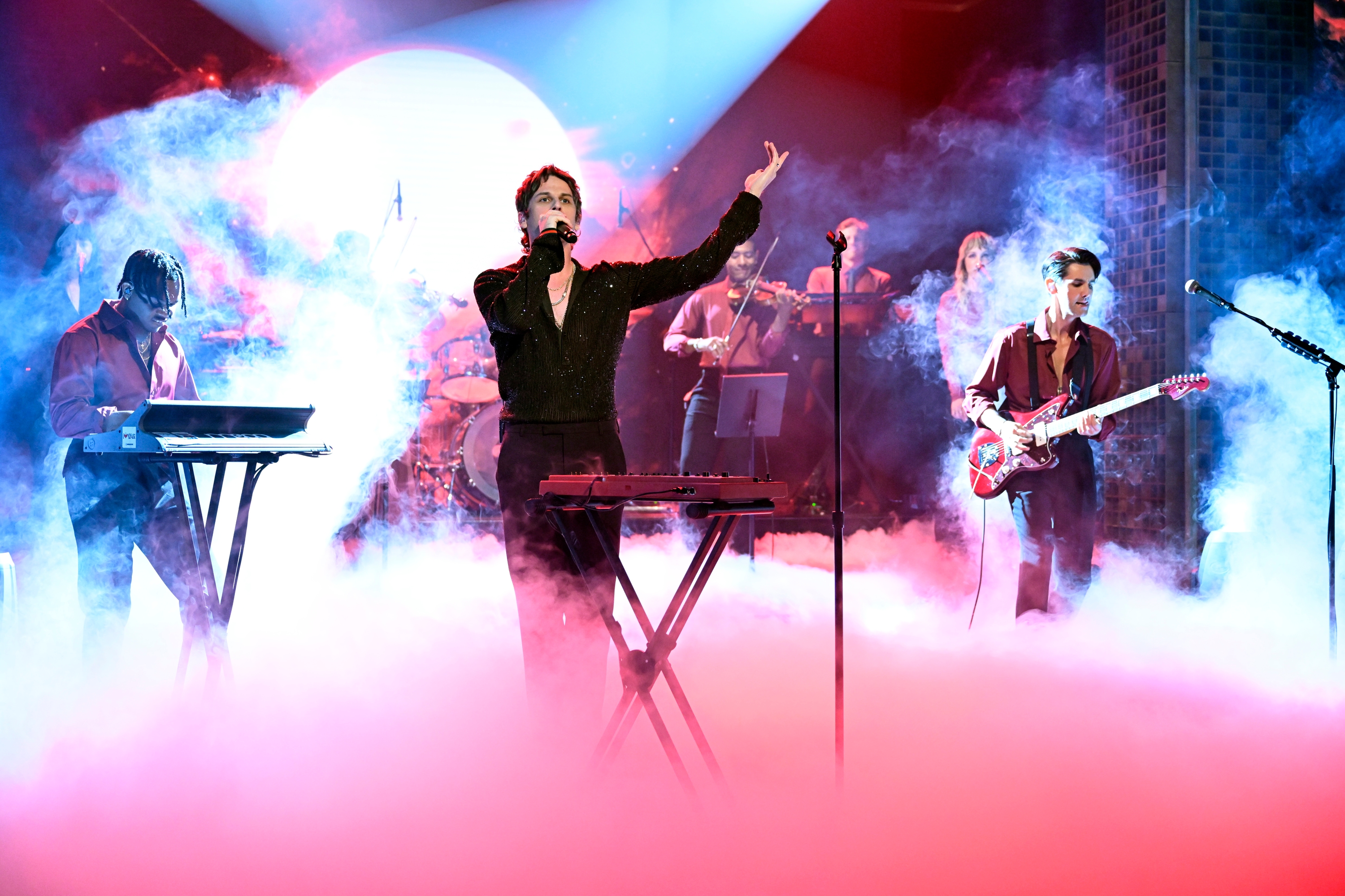 Band performing on stage surrounded by smoke, with lead singer at a keyboard. Other members play instruments like guitar and keyboard